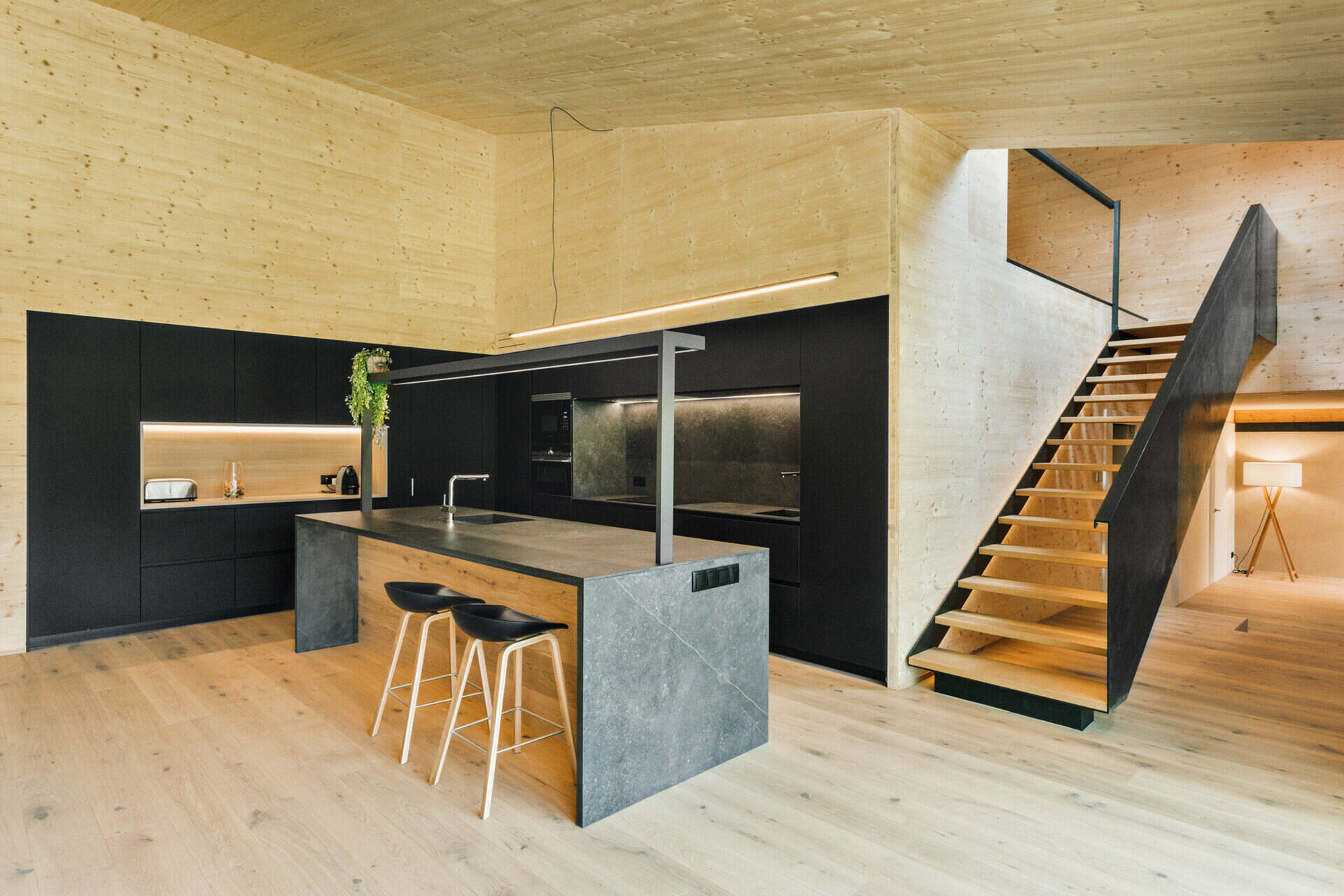 Black kitchen cabinetry and a grey island sit beside a dining bench backed by a timber pony wall.
