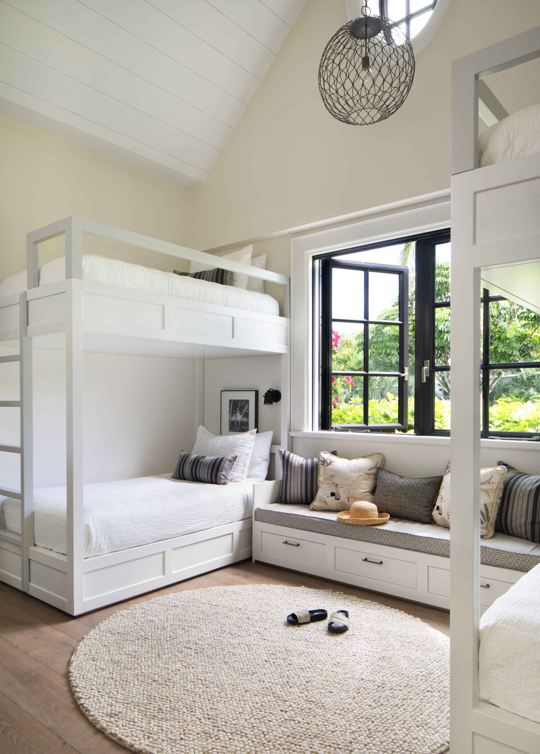 A charming bunk room feels bright, breezy, and ready for guests of any age. Crisp white built-in bunks and shiplap ceilings keep the space clean and classic, while a cozy window seat lined with striped and patterned pillows creates the perfect reading nook.