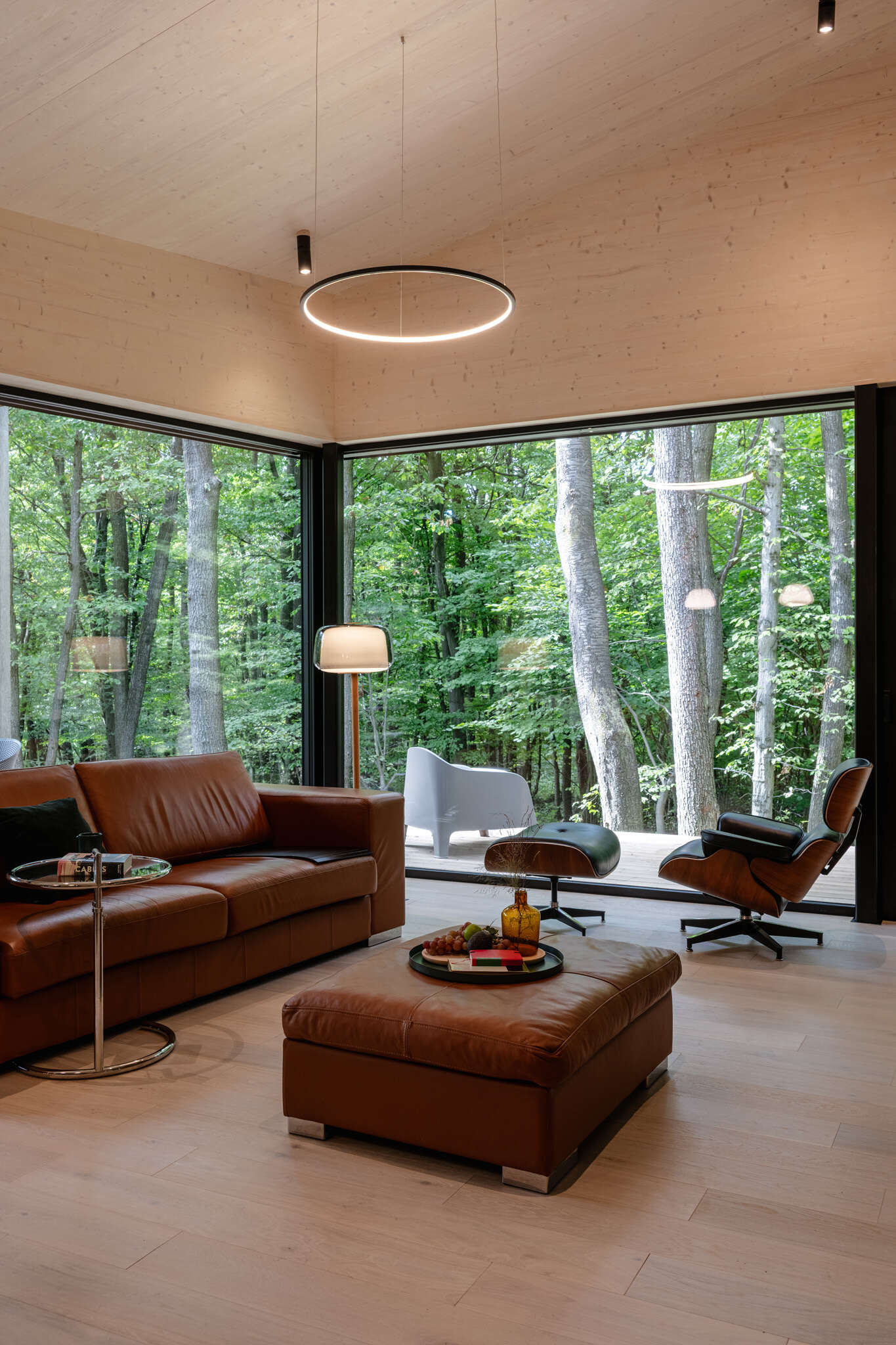 The living room opens toward the surrounding forest through large glass windows. Clean lines and exposed timber define the space, allowing light and landscape to take center stage, while designer furniture and premium materials add comfort without overwhelming the architecture. A full wall of shelving with a rolling ladders surrounds the TV and creates visual for the room.