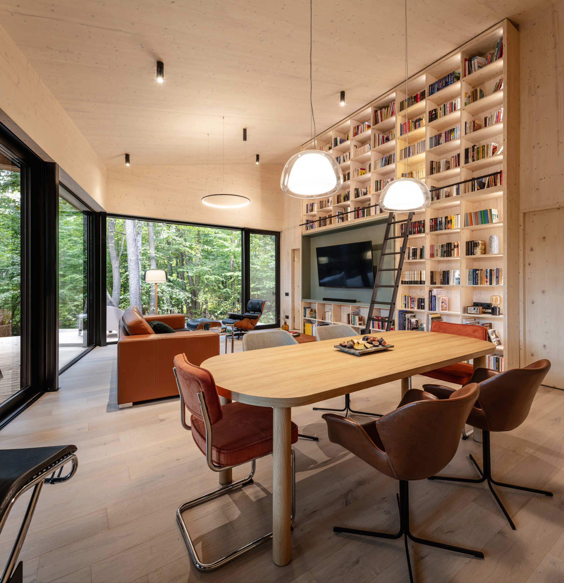 The living room opens toward the surrounding forest through large glass windows. Clean lines and exposed timber define the space, allowing light and landscape to take center stage, while designer furniture and premium materials add comfort without overwhelming the architecture. A full wall of shelving with a rolling ladders surrounds the TV and creates visual for the room.