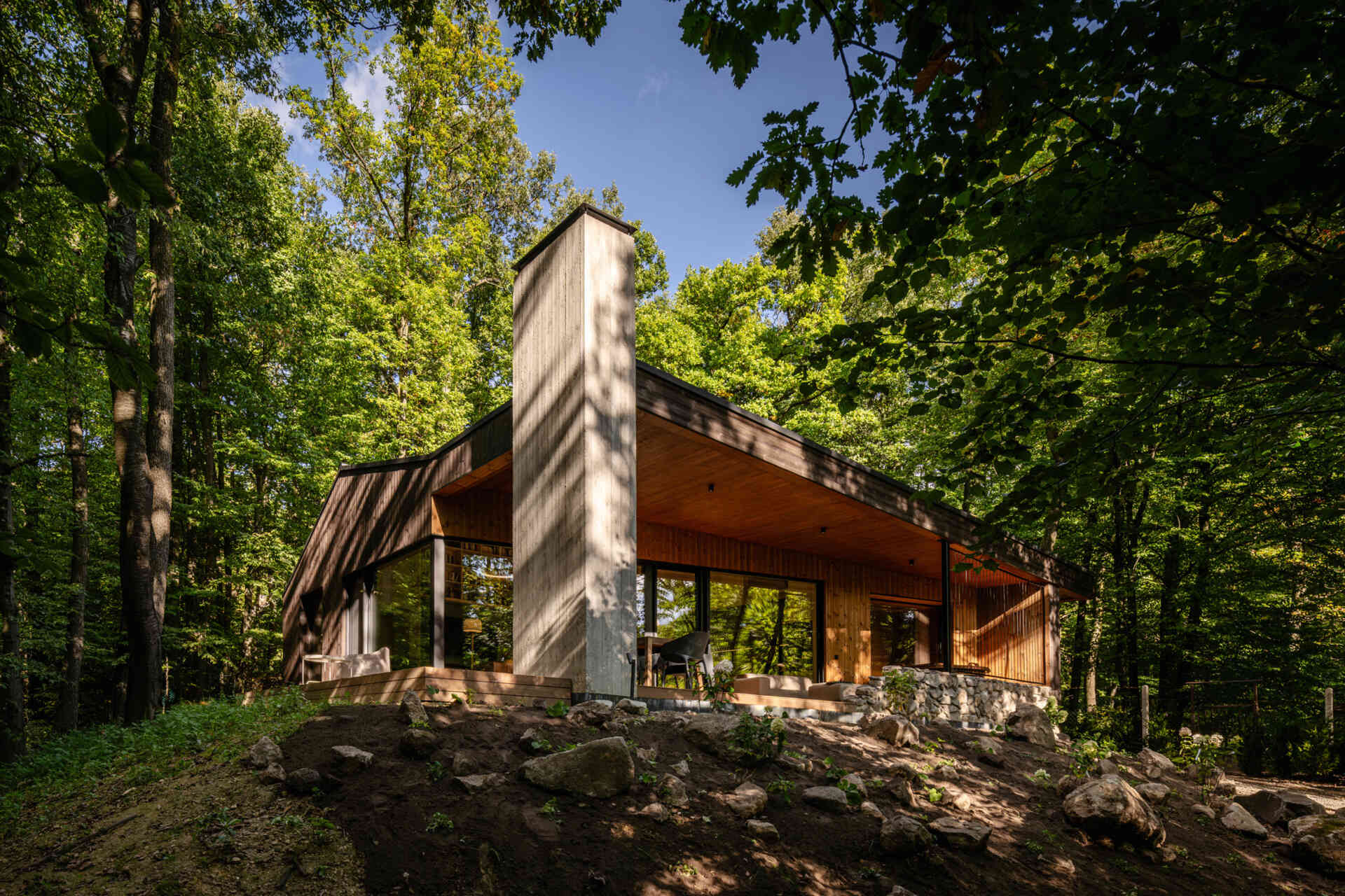 A modern forest home built from CLT and concrete, defined by a sculptural chimney and large glass openings.