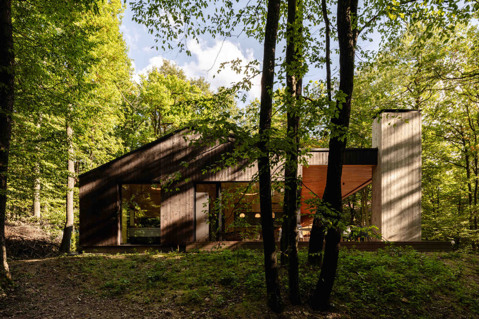 A modern forest home built from CLT and concrete, defined by a sculptural chimney and large glass openings.