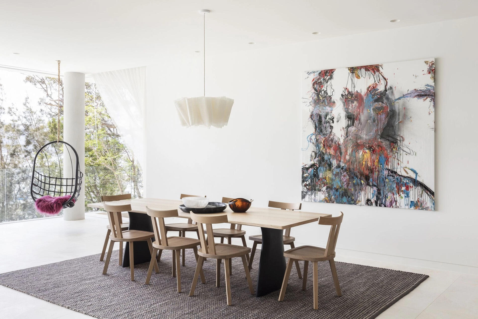The living room, dining area and kitchen share a continuous open plan space. A large rug and single pendant light anchor the dining table, defining the space without blocking views or flow. The layout encourages casual interaction and allows for entertaining, with smooth transitions to outdoor terraces and the garden below.