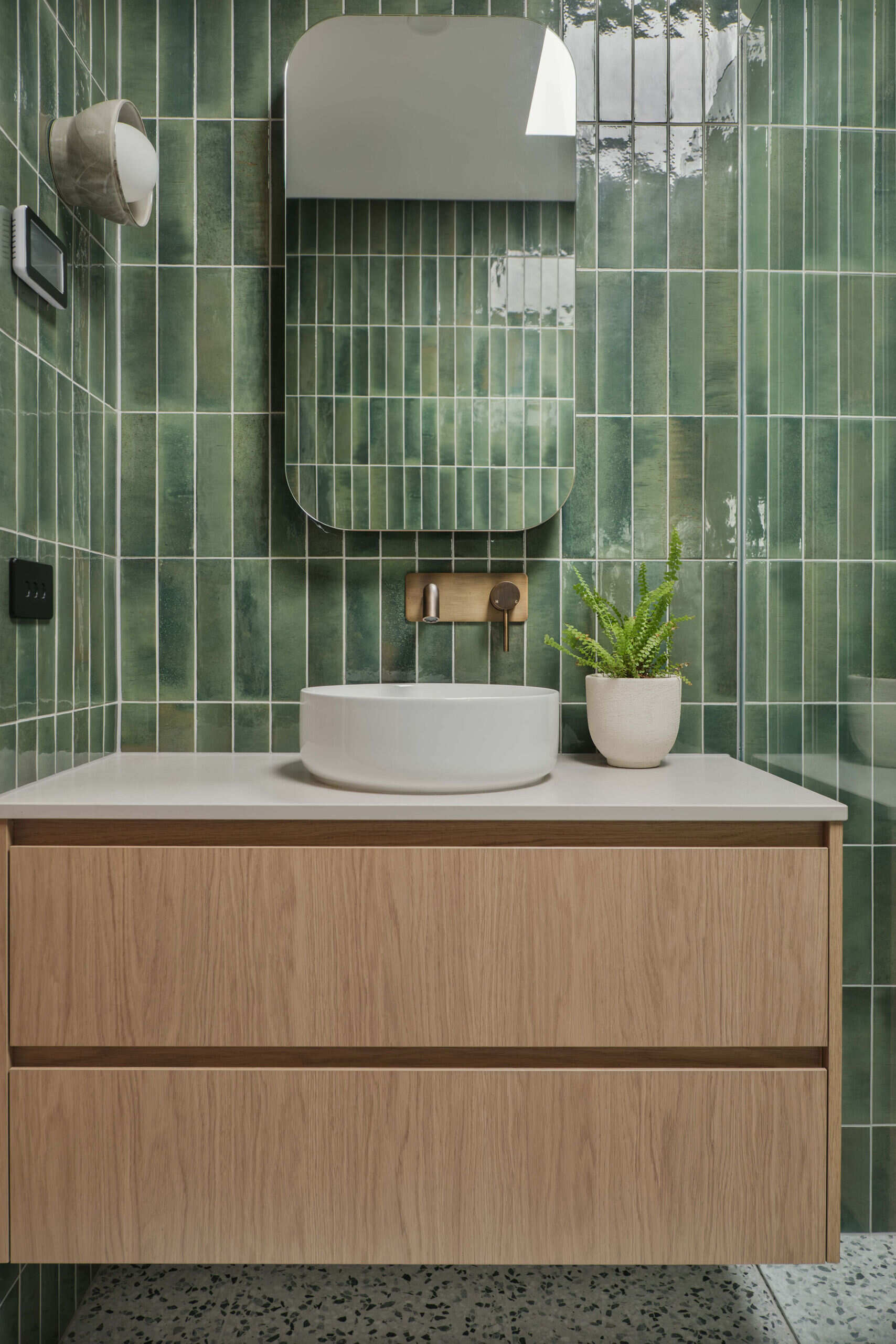 This guest bathroom features Gloss Mojave green tiles, Boston Oak joinery and terrazzo flooring create a warm and welcoming palette. A Smartstone bench top sits beneath a round white basin, and brushed vintage brass fittings add character. The shaving cabinet reflects the space beautifully, giving the room a fresh, polished feel.