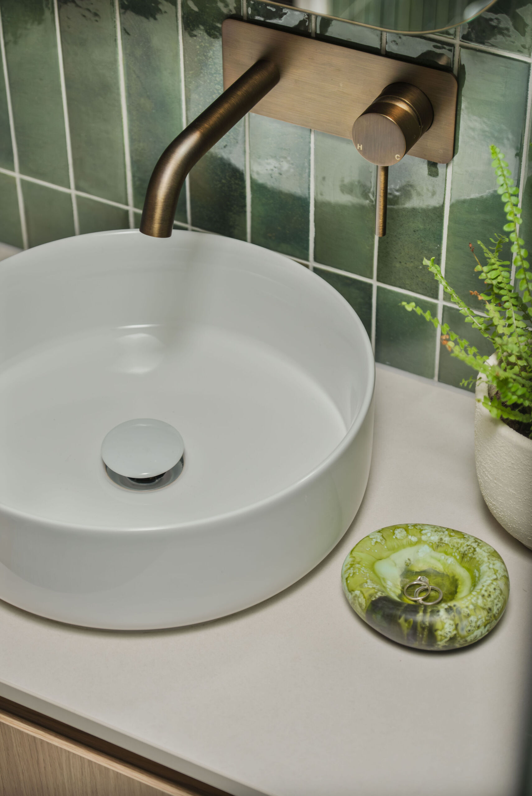This guest bathroom features Gloss Mojave green tiles, Boston Oak joinery and terrazzo flooring create a warm and welcoming palette. A Smartstone bench top sits beneath a round white basin, and brushed vintage brass fittings add character. The shaving cabinet reflects the space beautifully, giving the room a fresh, polished feel.