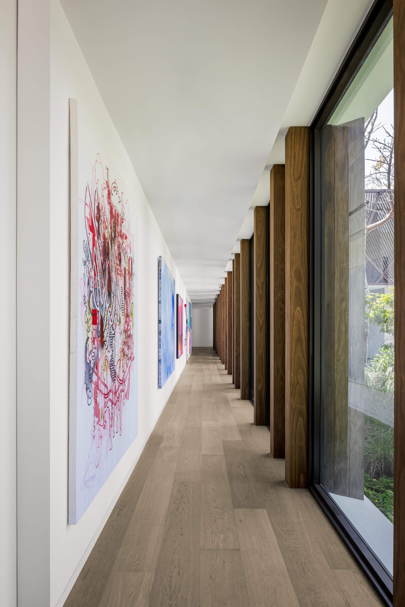This hallway feels like a bright, modern art walk, with clean white walls that let the colorful pieces stand out. Warm wood columns and big windows line one side, bringing in dappled daylight and glimpses of the garden as you move through the space.