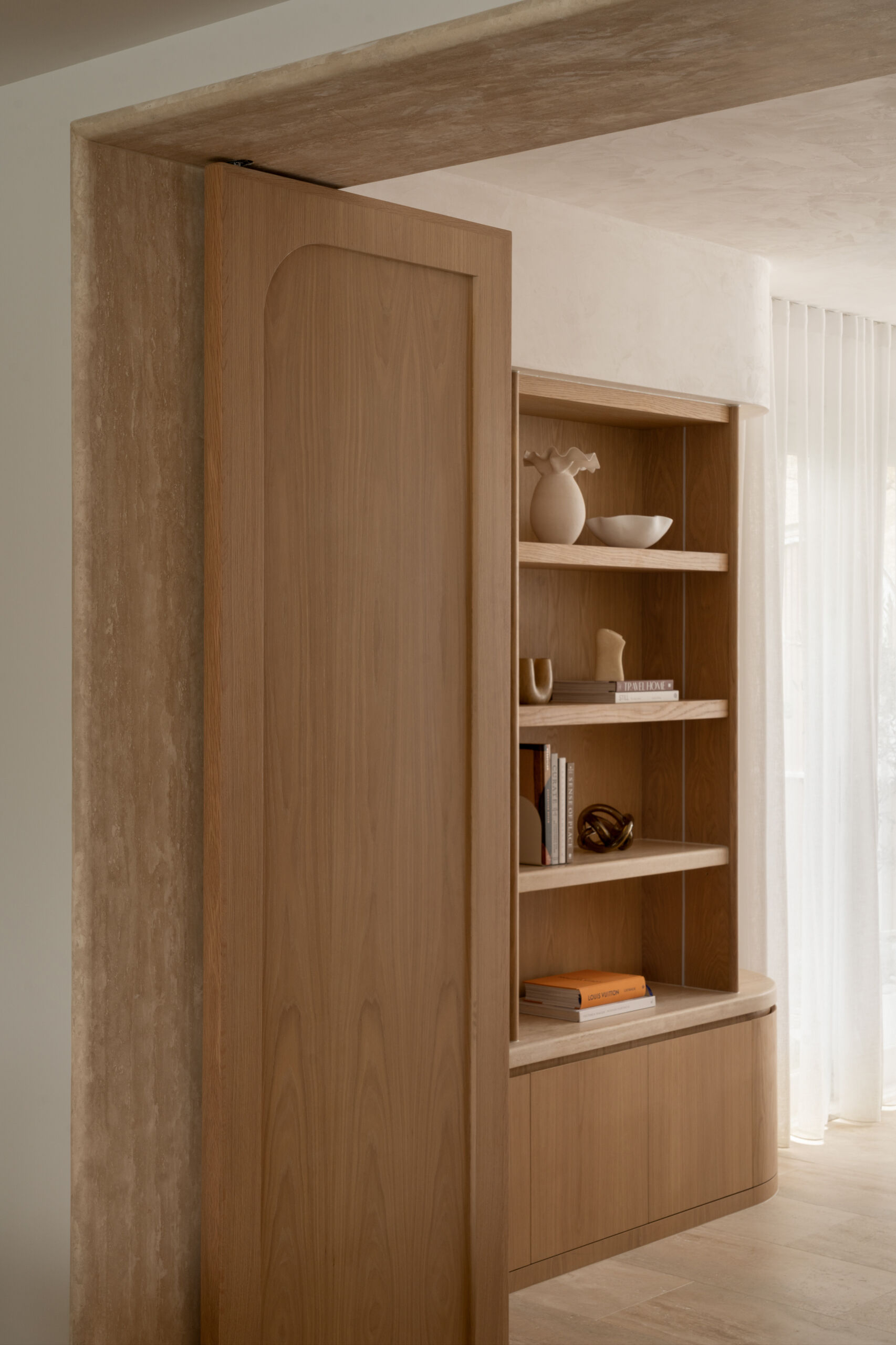 A sophisticated home office with custom oak doors, curved shelving, a travertine threshold, and soft Venetian-plaster light.