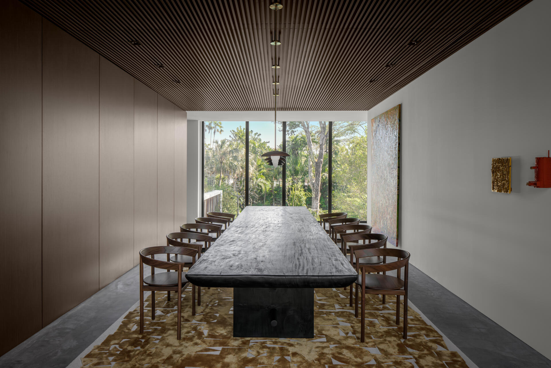 This dining room feels like a modern gallery, warmed up with rich wood and quiet texture. A long, substantial table anchors the space, paired with sleek wood chairs that keep the lines clean but still inviting. The slatted wood ceiling adds depth overhead, while the oversized artwork brings just enough color to soften the minimal palette.