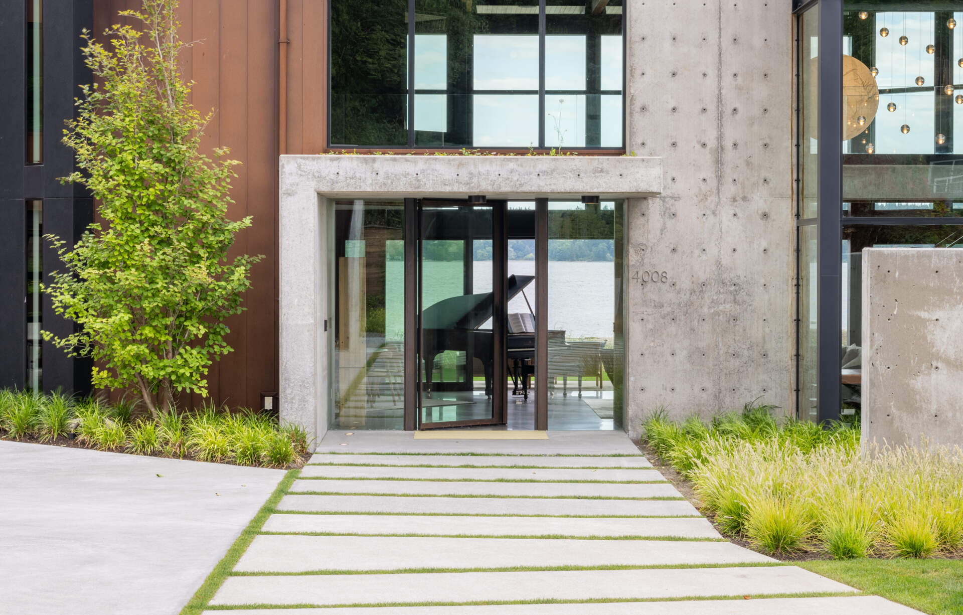 The entry is defined by exposed architectural concrete, immediately establishing the home’s material language. Above the entry sits a green roof, which becomes visible from inside the house. This moment connects the exterior landscape to the interior circulation, while a pivoting glass door welcomes visitors.