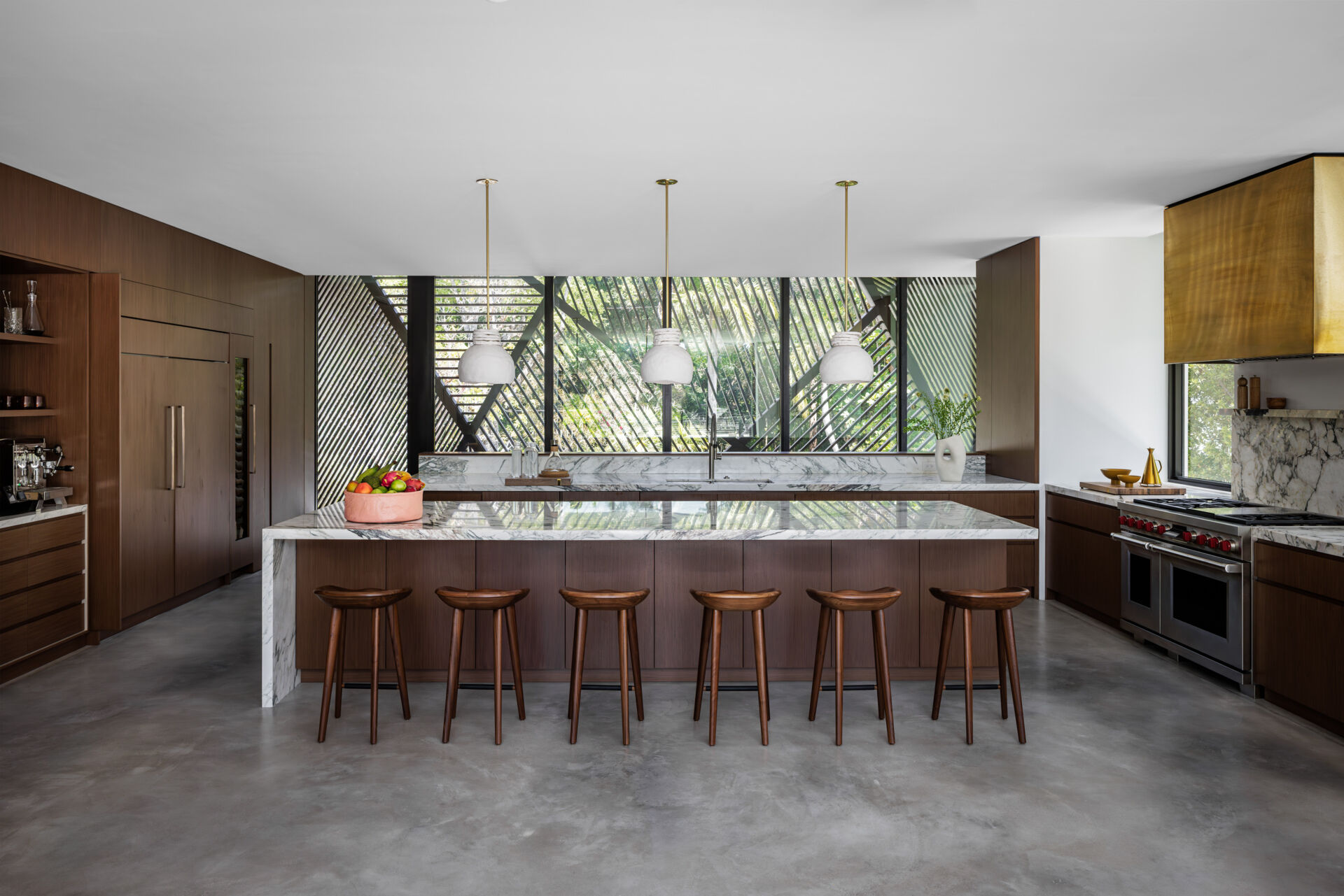 This modern kitchen feels grounded and uncluttered, with deep wood cabinets, a smooth concrete floor, and a marble island at its center. Simple pendant lights hang above, casting soft light over the counter and warm wood stools. Wide windows frame leafy views, while a brass range hood adds a metallic accent.