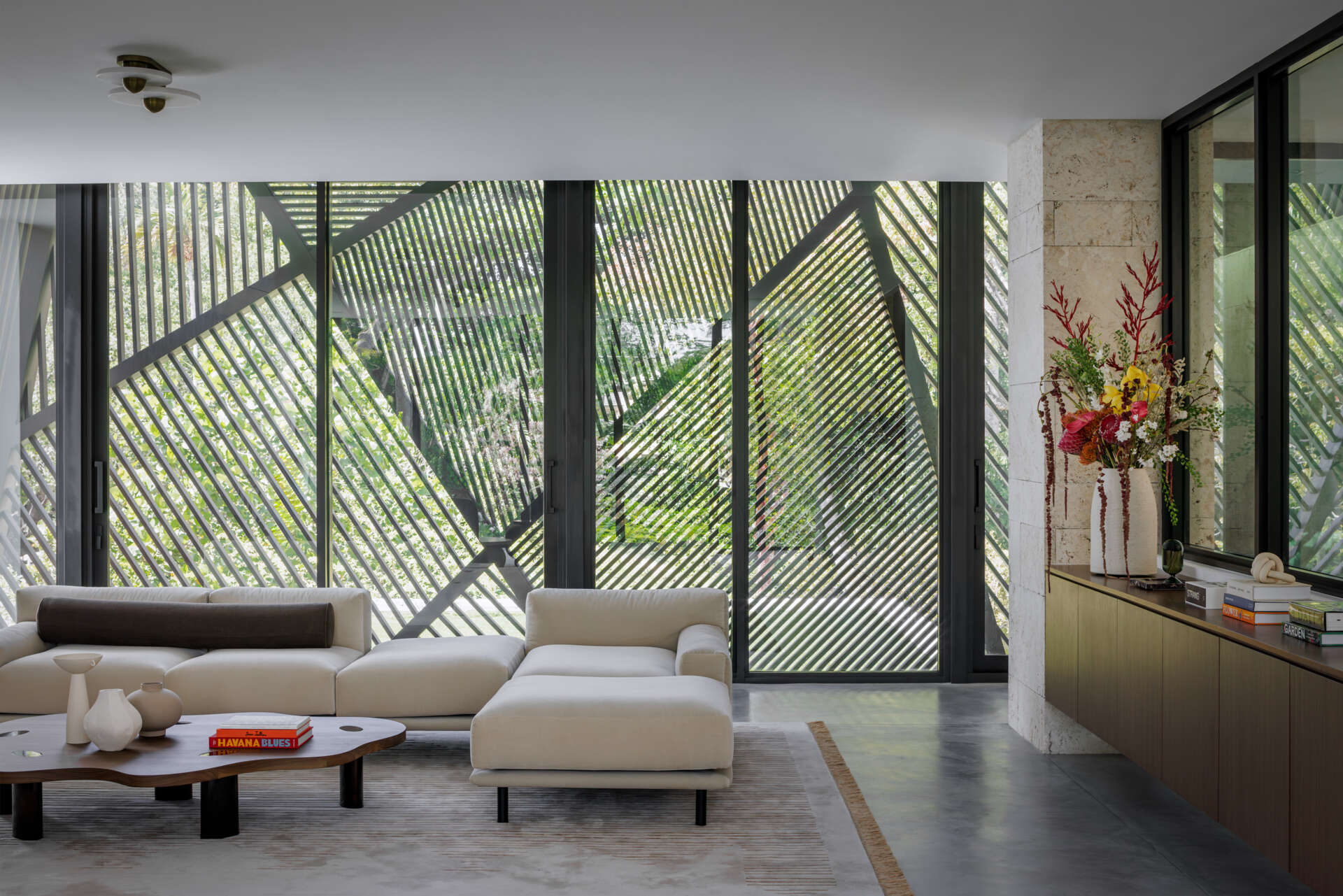 This living room feels light and composed, with creamy walls and a concrete floor that highlights the furniture’s forms. Curved sofas and plush chairs in soft caramel and cinnamon tones bring warmth, while layered rugs, wood elements, and sculptural side tables add subtle texture, and oversized artwork adds subtle color.