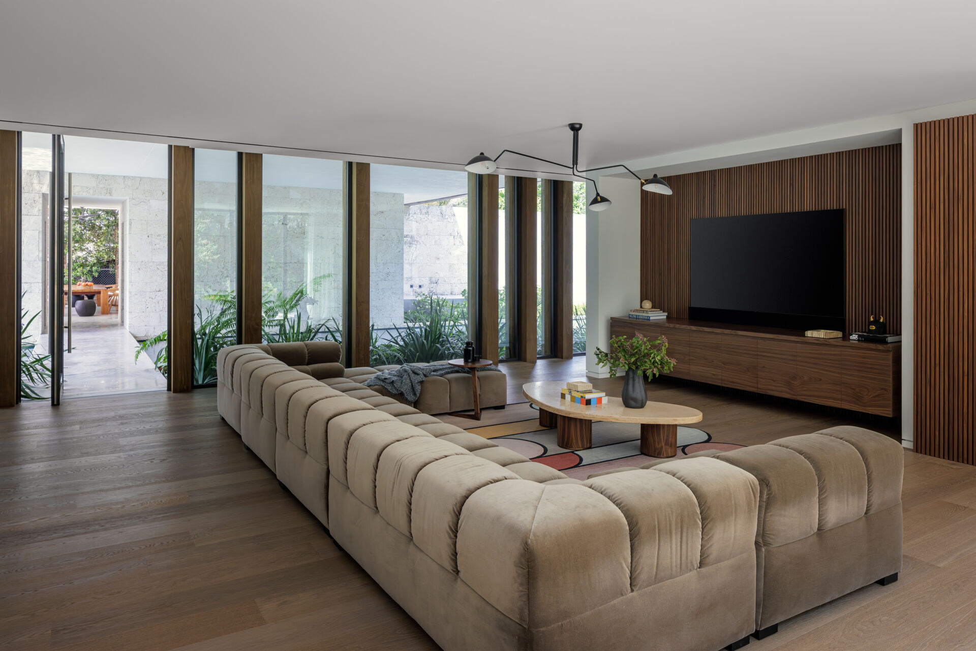 This modern media room feels calm and comfortable, with tufted seating in soft taupe and warm wood floors. A slatted wood wall and a long floating console keep the space clean and organized. Tall windows bring in garden views and natural light, while a simple sculptural ceiling fixture adds a modern accent.
