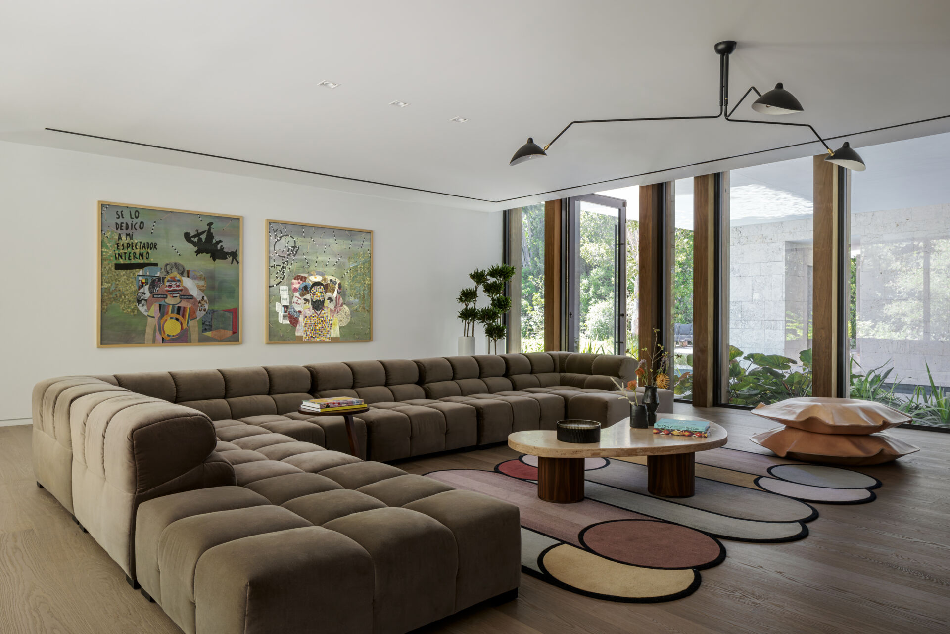 This modern media room feels calm and comfortable, with tufted seating in soft taupe and warm wood floors. A slatted wood wall and a long floating console keep the space clean and organized. Tall windows bring in garden views and natural light, while a simple sculptural ceiling fixture adds a modern accent.