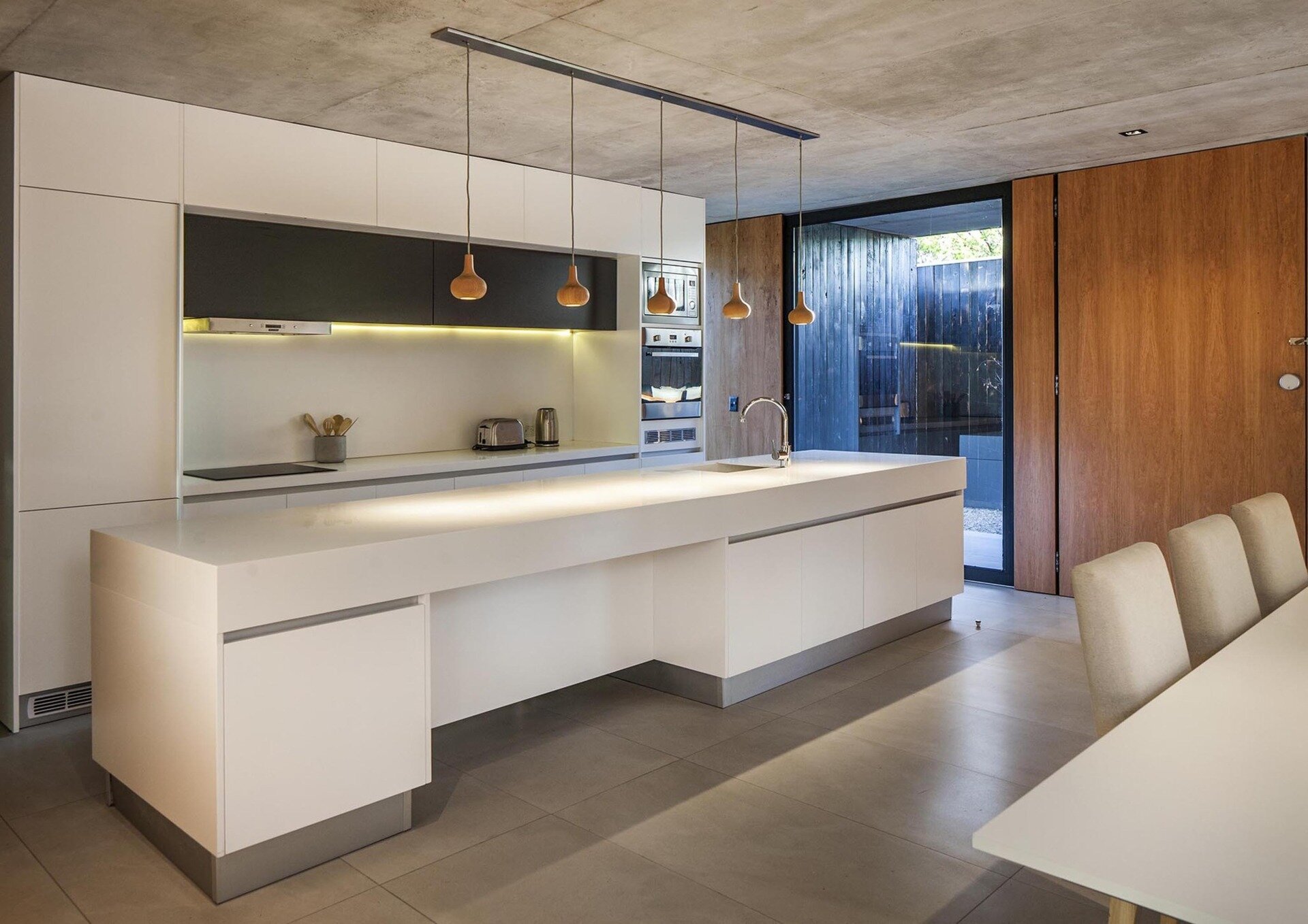 A minimalist white kitchen with a large island and wood pendant lights flows directly into the dining and living spaces.