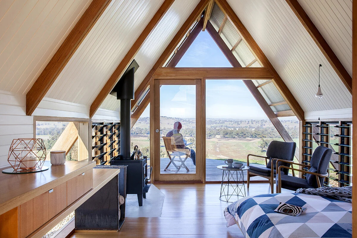 Inside this rural A-frame cabin, the windows follow the structural lines of the hut, drawing light into the small living area. A black freestanding wood burner keeps the space warm on chilly nights, while louvre windows allow fresh air to flow on warmer days. A deck extends from the main living area, providing a front-row seat to the valley views.
