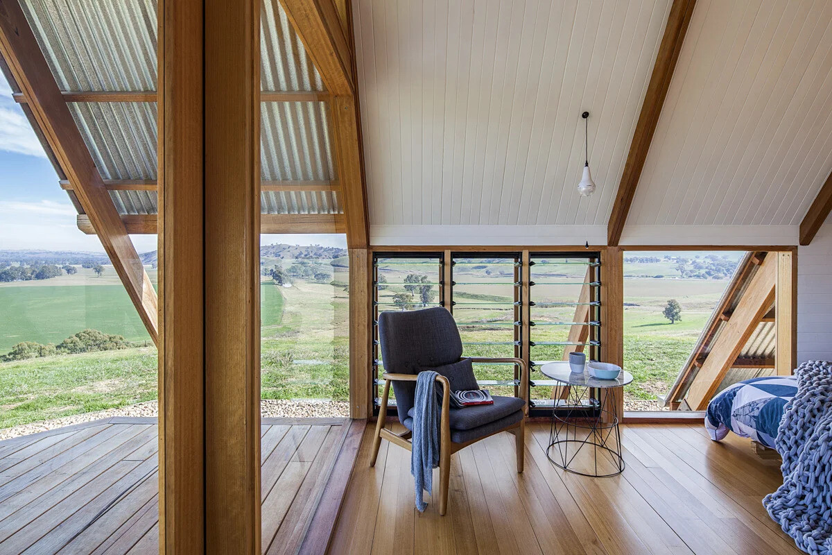 Inside this rural A-frame cabin, the windows follow the structural lines of the hut, drawing light into the small living area. A black freestanding wood burner keeps the space warm on chilly nights, while louvre windows allow fresh air to flow on warmer days. A deck extends from the main living area, providing a front-row seat to the valley views.
