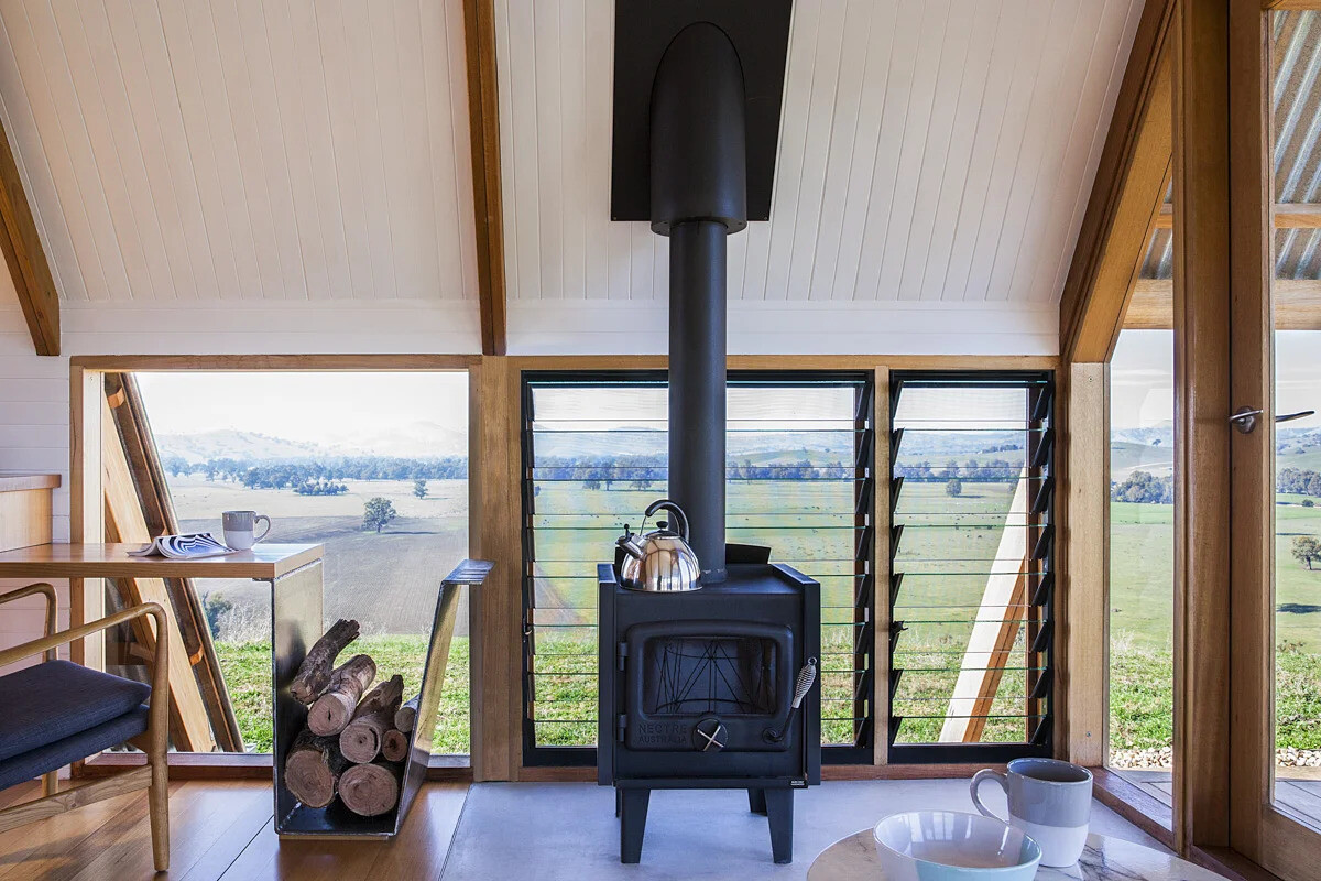 Inside this rural A-frame cabin, the windows follow the structural lines of the hut, drawing light into the small living area. A black freestanding wood burner keeps the space warm on chilly nights, while louvre windows allow fresh air to flow on warmer days. A deck extends from the main living area, providing a front-row seat to the valley views.