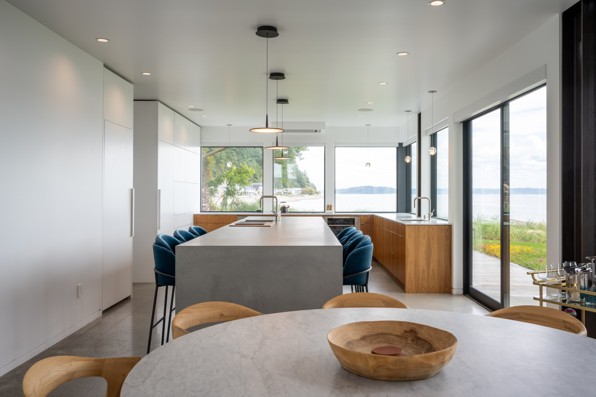 The kitchen has a long concrete island anchors the space, providing room for everyday use and casual gathering. Minimalist white cabinetry is paired with warm wood base units, keeping the palette restrained while adding subtle contrast. Floor to ceiling windows line the perimeter, drawing in natural light and uninterrupted water views. 