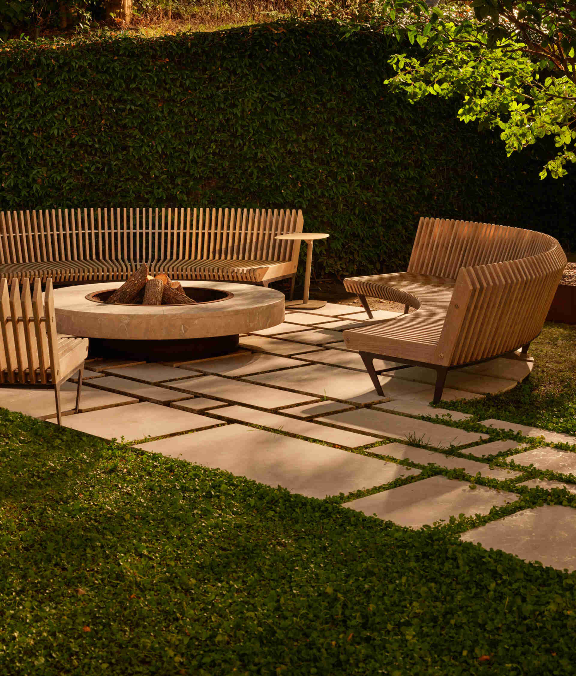Tucked in a lush corner, the firepit lounge invites slow evenings and conversation. A curved wood-slat bench wraps around a smooth stone fire bowl. Large pavers with grassy seams keep the area tidy, while surrounding greenery adds privacy and dappled shade during the day.