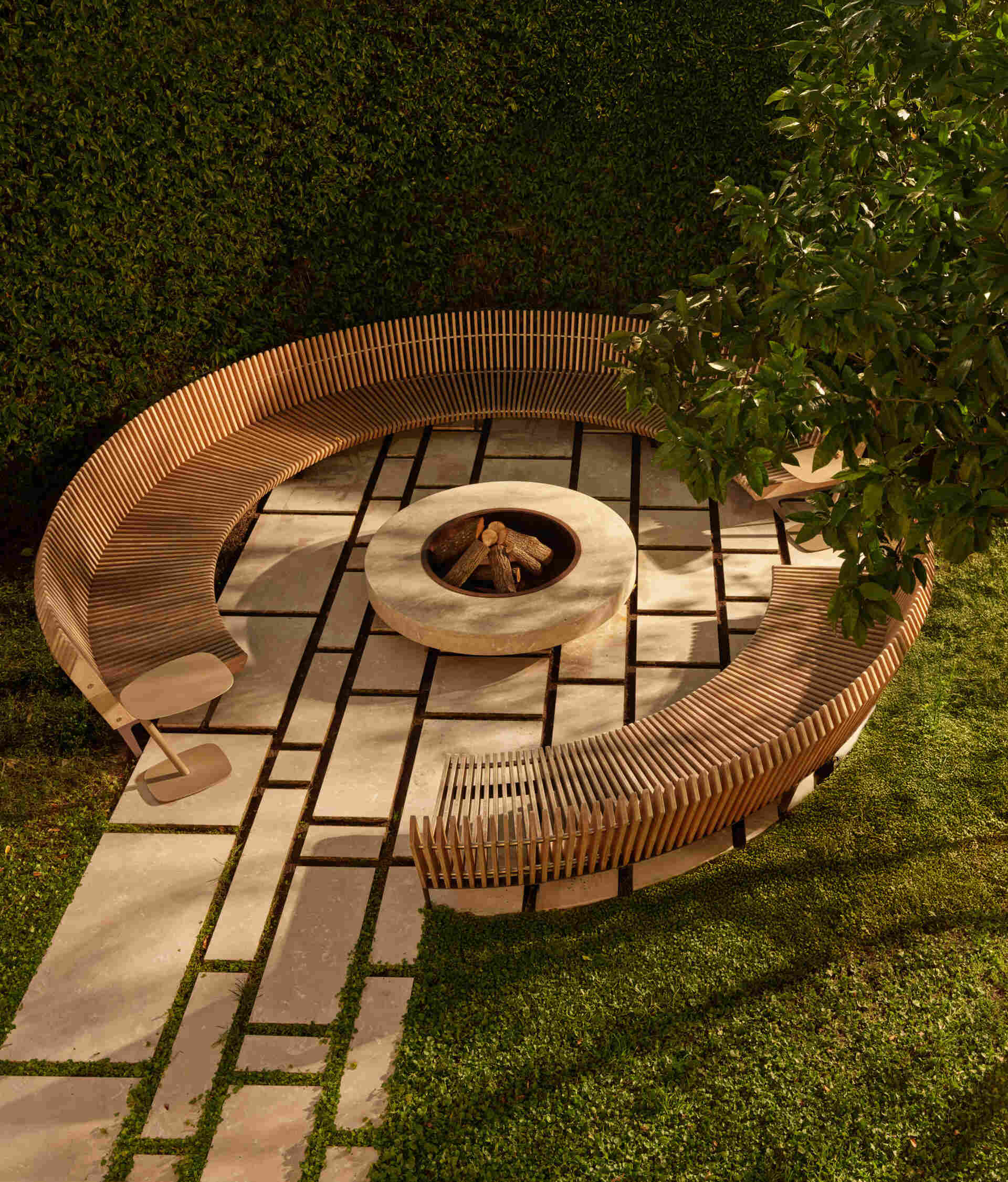 Tucked in a lush corner, the firepit lounge invites slow evenings and conversation. A curved wood-slat bench wraps around a smooth stone fire bowl. Large pavers with grassy seams keep the area tidy, while surrounding greenery adds privacy and dappled shade during the day.