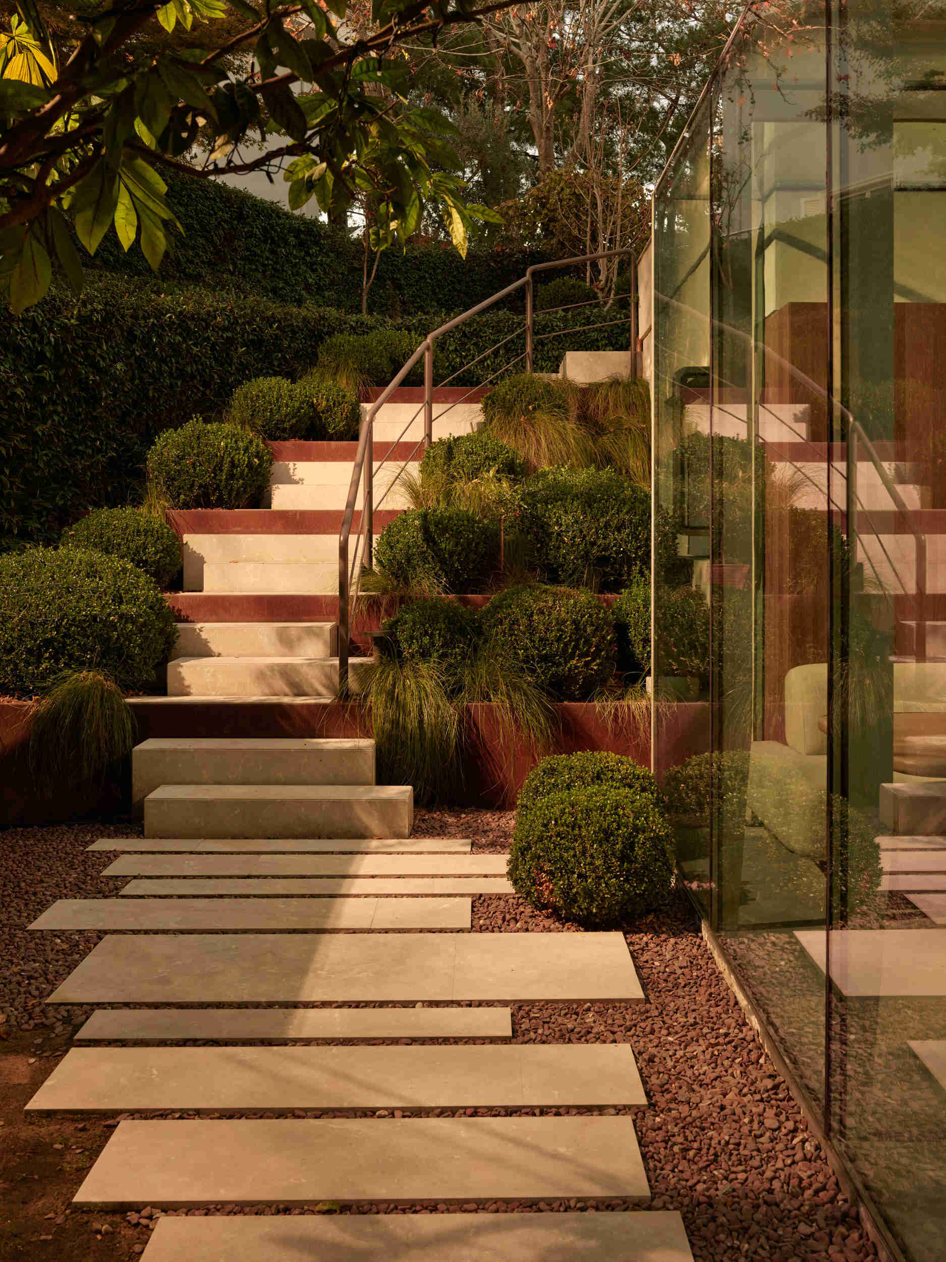 Garden stairs feel like a walk through a living sculpture. Pale stepping stones float over warm gravel, with tiered concrete steps rising between rust-colored planters. Ornamental grasses sway gently, and a slim metal handrail adds a modern line.