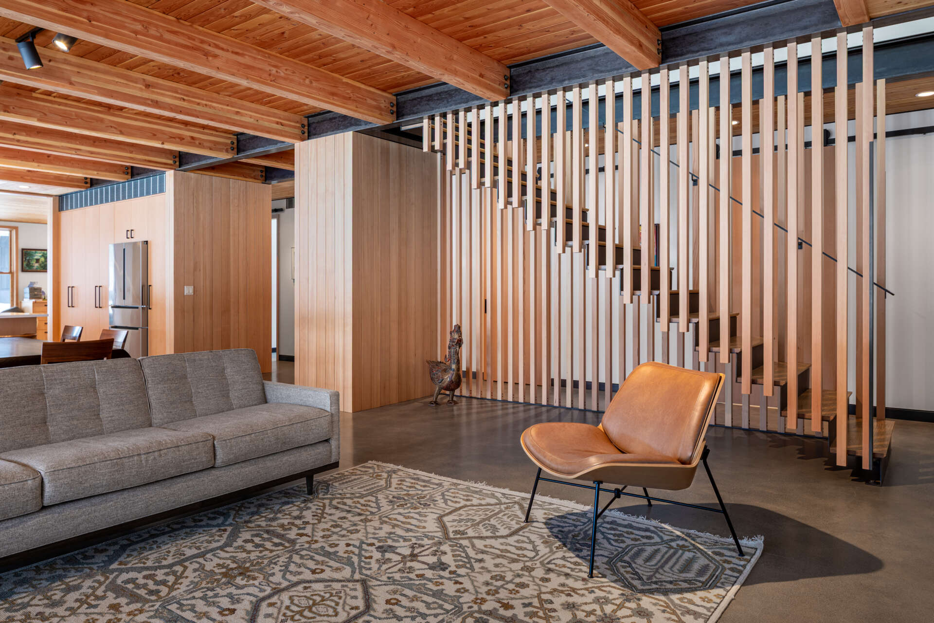 An open living and dining space warmed by wood ceilings and beams, with concrete floors, soft furnishings, and wide glass doors framing snowy valley views.