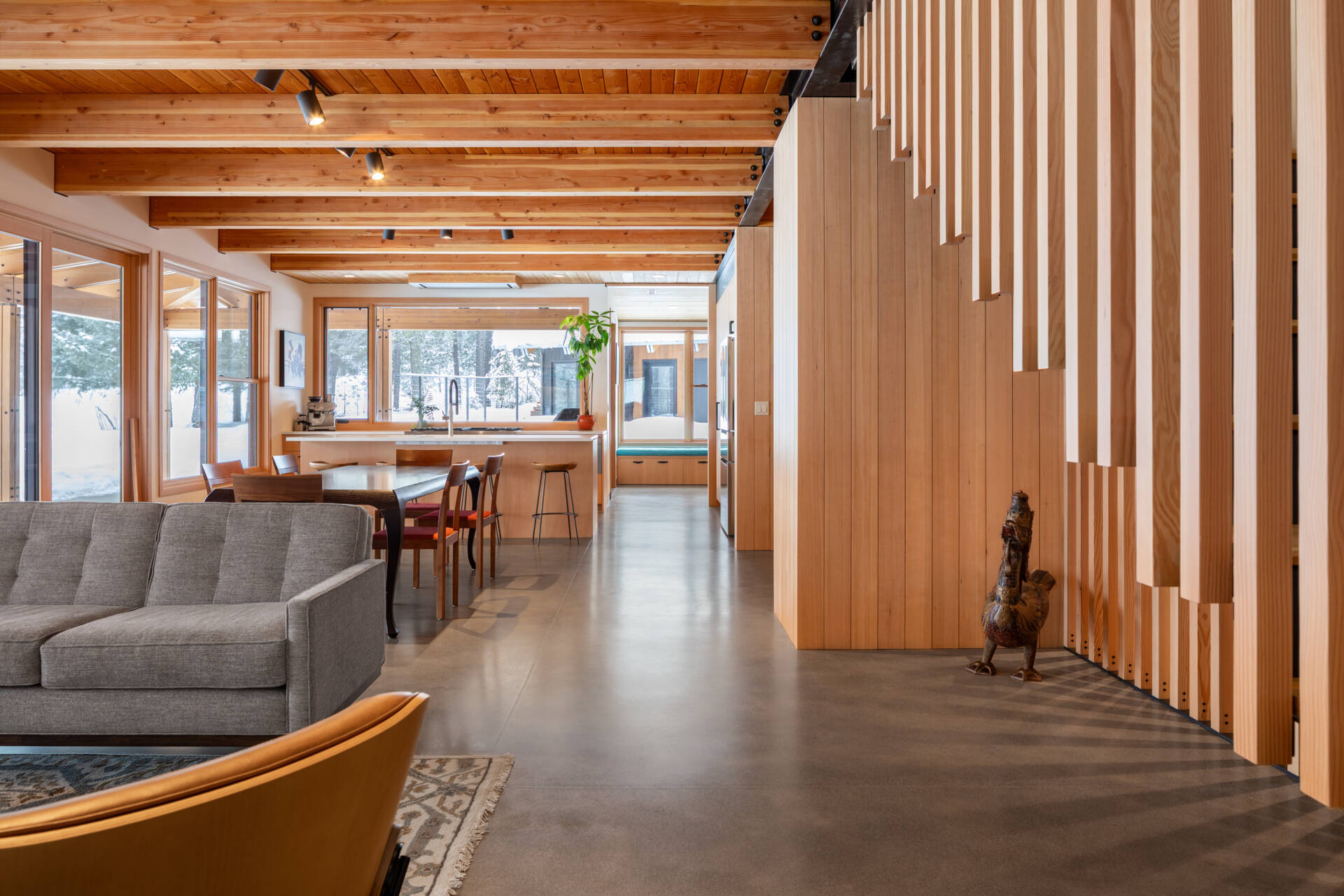 An open living and dining space warmed by wood ceilings and beams, with concrete floors, soft furnishings, and wide glass doors framing snowy valley views.