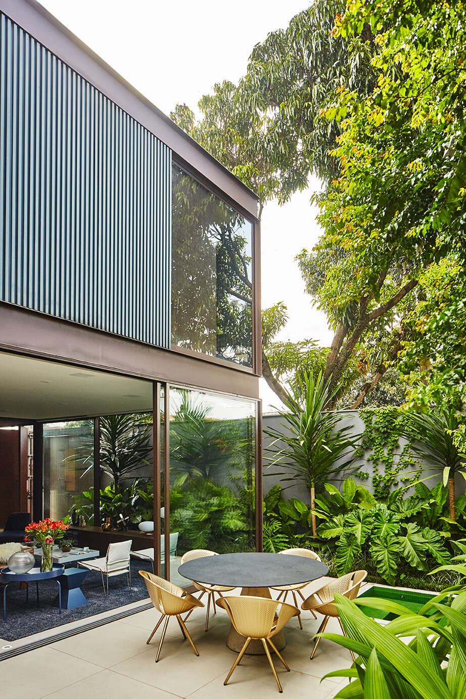 From the garden, the second floor becomes a defining feature. The upper level appears to hover above the ground floor, resting on a transparent base of glass. This contrast between the heavy opaque volume above and the lightness below gives the house its distinctive floating effect. Tropical landscaping surrounds the space, softening the concrete walls and bringing greenery deep into the home.