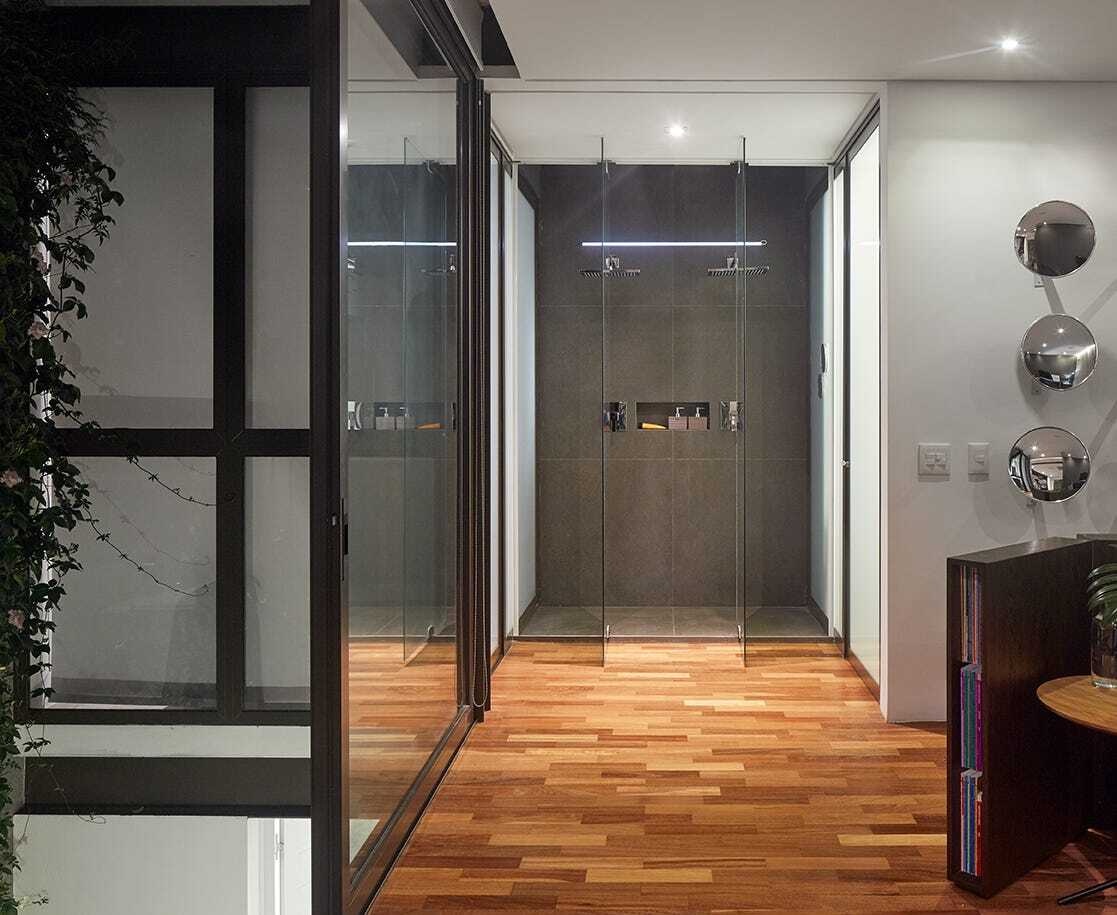Behind the bed, the ensuite bathroom unfolds. A glass enclosed shower becomes the focal point, while white doors on either side lead separately to the washroom and a walk in closet. The arrangement maintains privacy while preserving a sense of openness within the bedroom.