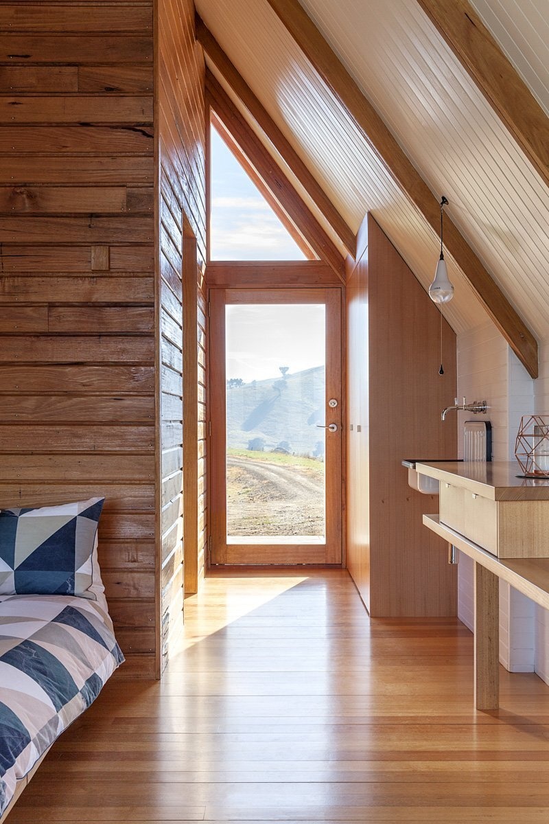 Inside this rural A-frame cabin, the windows follow the structural lines of the hut, drawing light into the small living area. A black freestanding wood burner keeps the space warm on chilly nights, while louvre windows allow fresh air to flow on warmer days. A deck extends from the main living area, providing a front-row seat to the valley views.