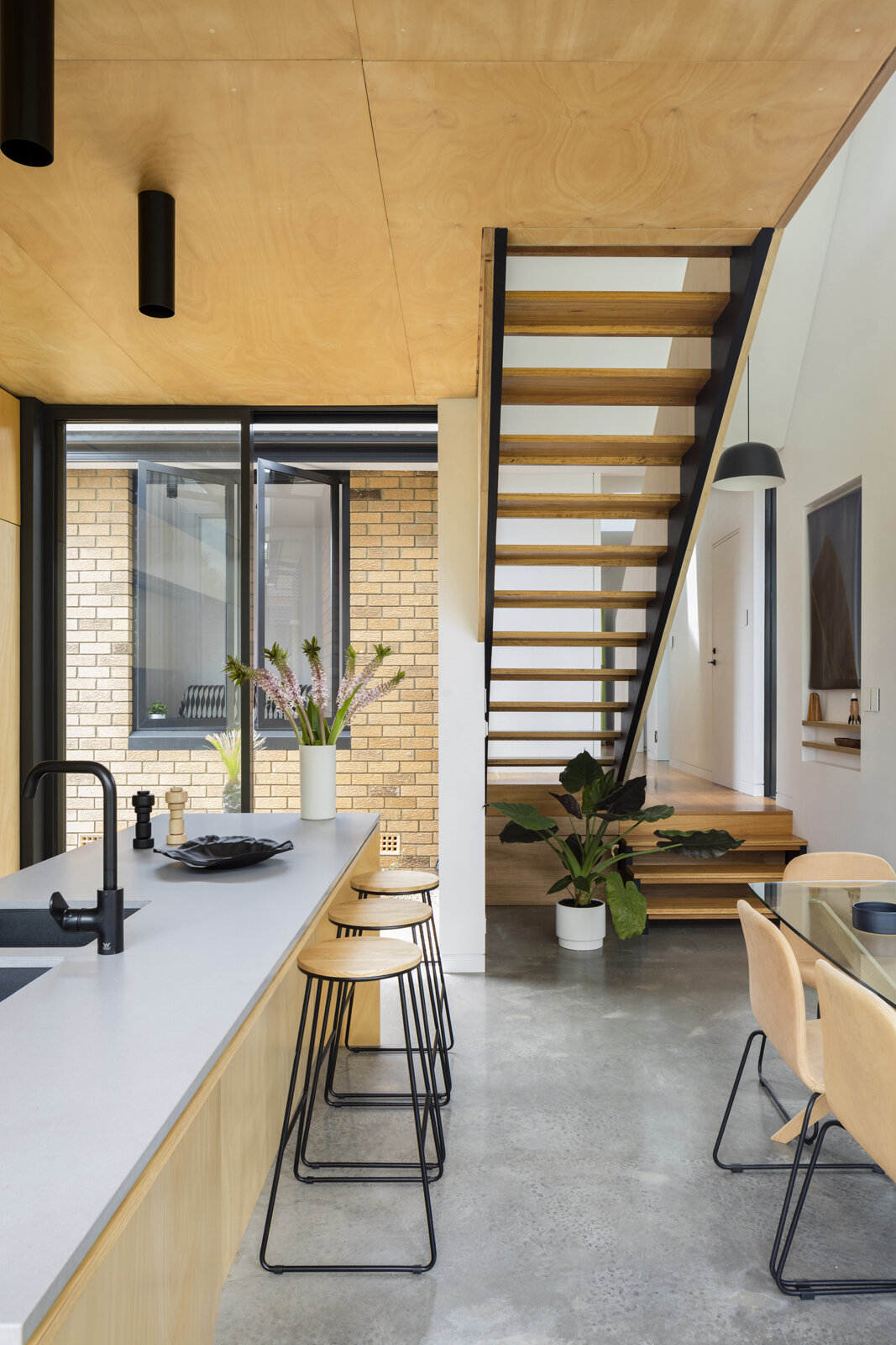 Off the hallway, a set of timber and steel stairs leads upward to a lofted area positioned above the kitchen. The stair continues the home’s material language while creating a clear vertical connection within the extension.