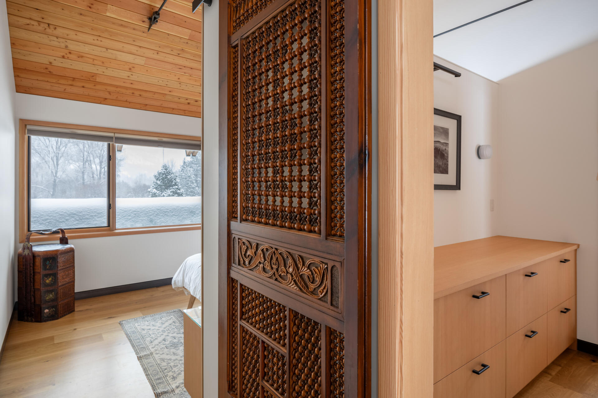 This primary suite feels serene and thoughtfully layered, mixing warm wood ceilings and pale built-ins with crisp white walls. A beautifully carved wood door adds a touch of old-world character, while the long dresser wall makes getting ready feel easy and uncluttered. In the bath, glossy white tile, a clean double vanity, and a sculptural soaking tub create a spa-like rhythm, softened by snowy views and gentle natural light.