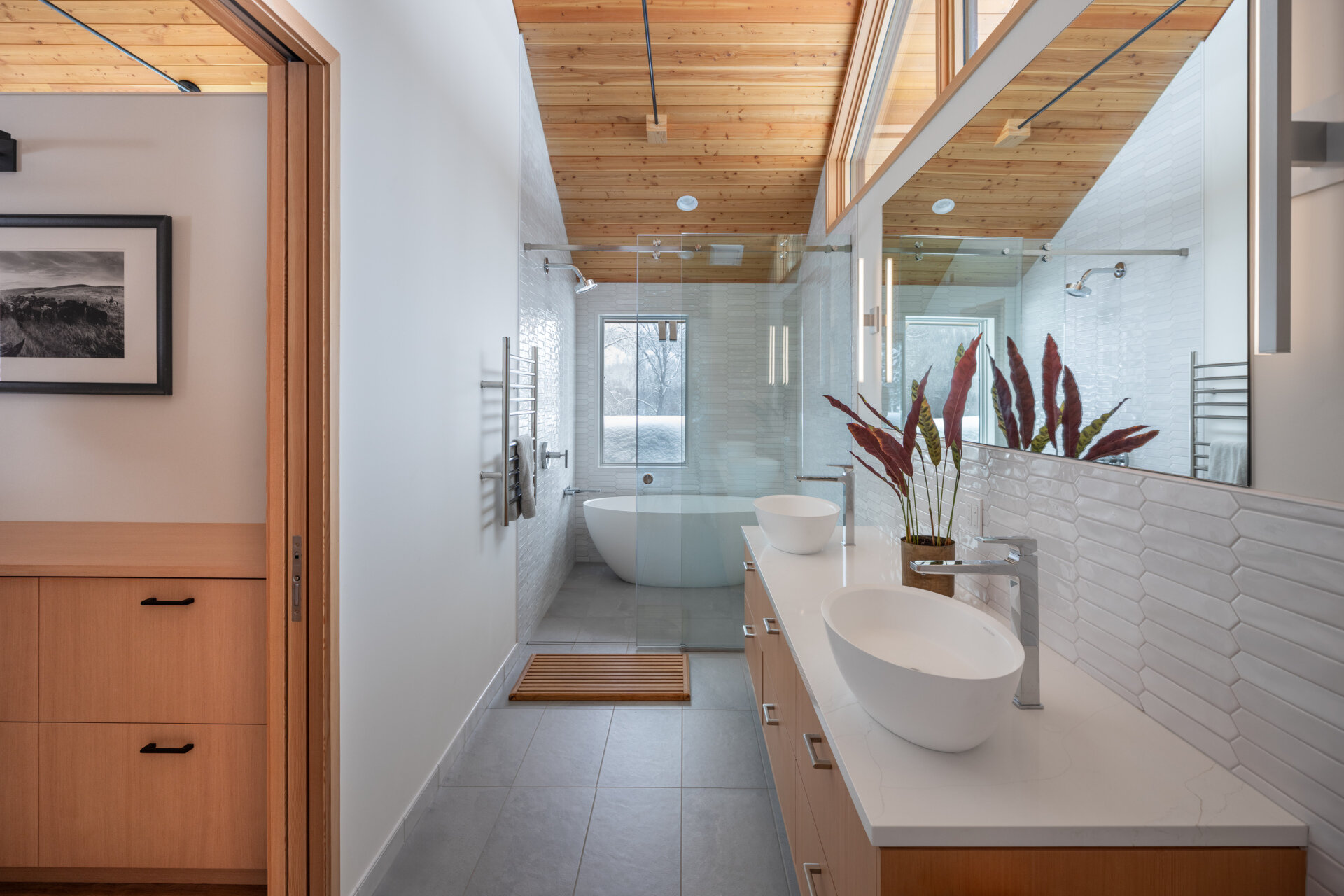 This primary suite feels serene and thoughtfully layered, mixing warm wood ceilings and pale built-ins with crisp white walls. A beautifully carved wood door adds a touch of old-world character, while the long dresser wall makes getting ready feel easy and uncluttered. In the bath, glossy white tile, a clean double vanity, and a sculptural soaking tub create a spa-like rhythm, softened by snowy views and gentle natural light.