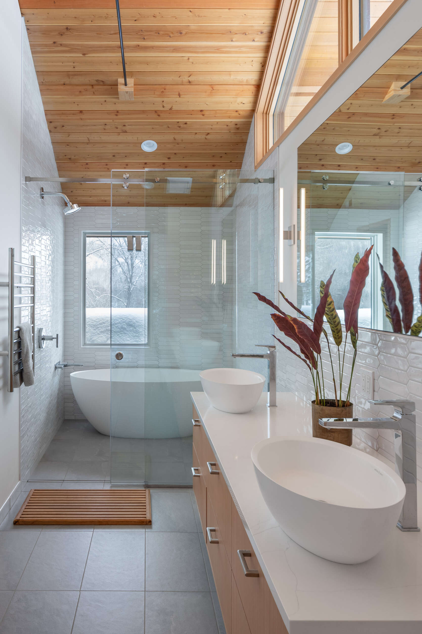 This primary suite feels serene and thoughtfully layered, mixing warm wood ceilings and pale built-ins with crisp white walls. A beautifully carved wood door adds a touch of old-world character, while the long dresser wall makes getting ready feel easy and uncluttered. In the bath, glossy white tile, a clean double vanity, and a sculptural soaking tub create a spa-like rhythm, softened by snowy views and gentle natural light.