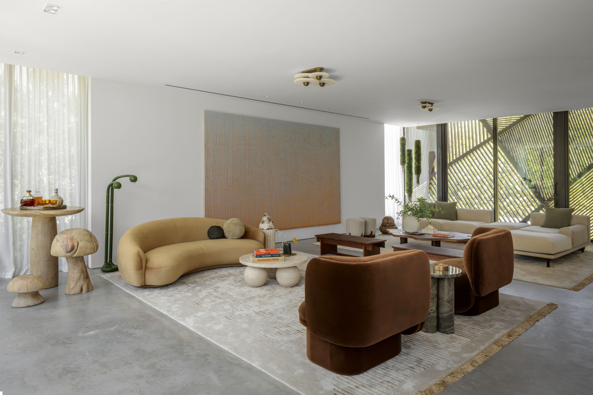 This living room feels light and composed, with creamy walls and a concrete floor that highlights the furniture’s forms. Curved sofas and plush chairs in soft caramel and cinnamon tones bring warmth, while layered rugs, wood elements, and sculptural side tables add subtle texture, and oversized artwork adds subtle color.