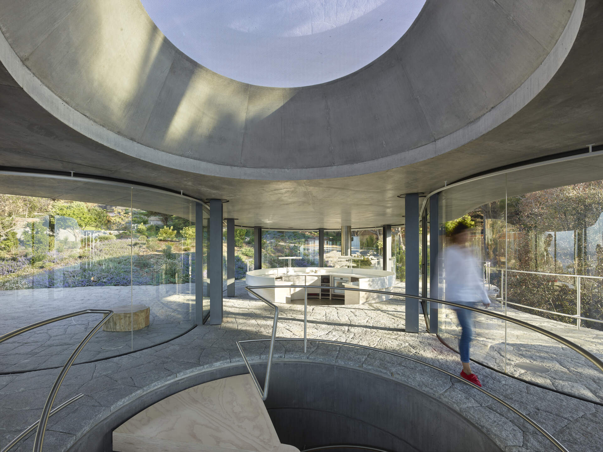 An open curved kitchen within the glass pavilion where structure fades and the coastal view becomes the focus.