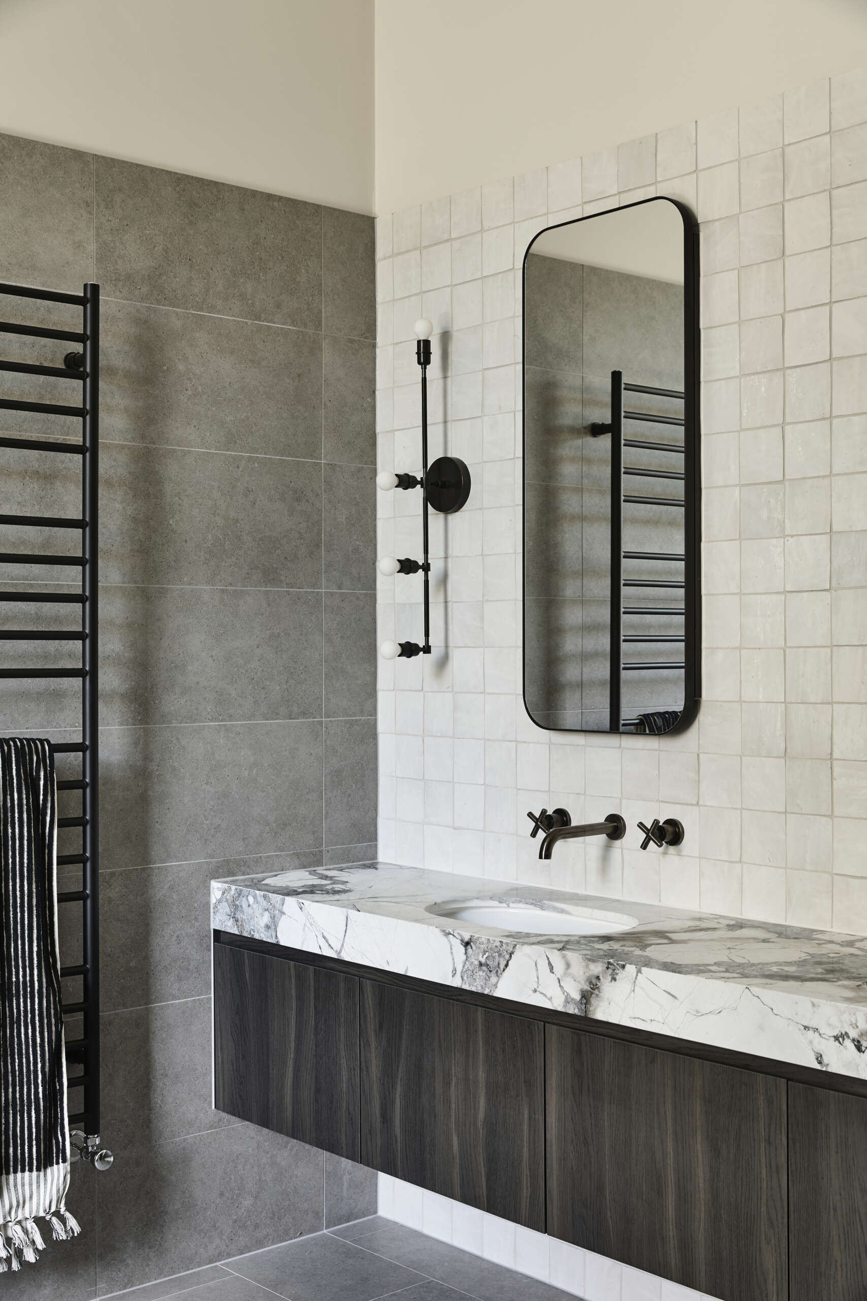 A calm and tailored bathroom wrapped in charcoal-gray tile with a floating dark wood vanity and white marble tops. Black fixtures and freestanding tubs create a spa-like feel.