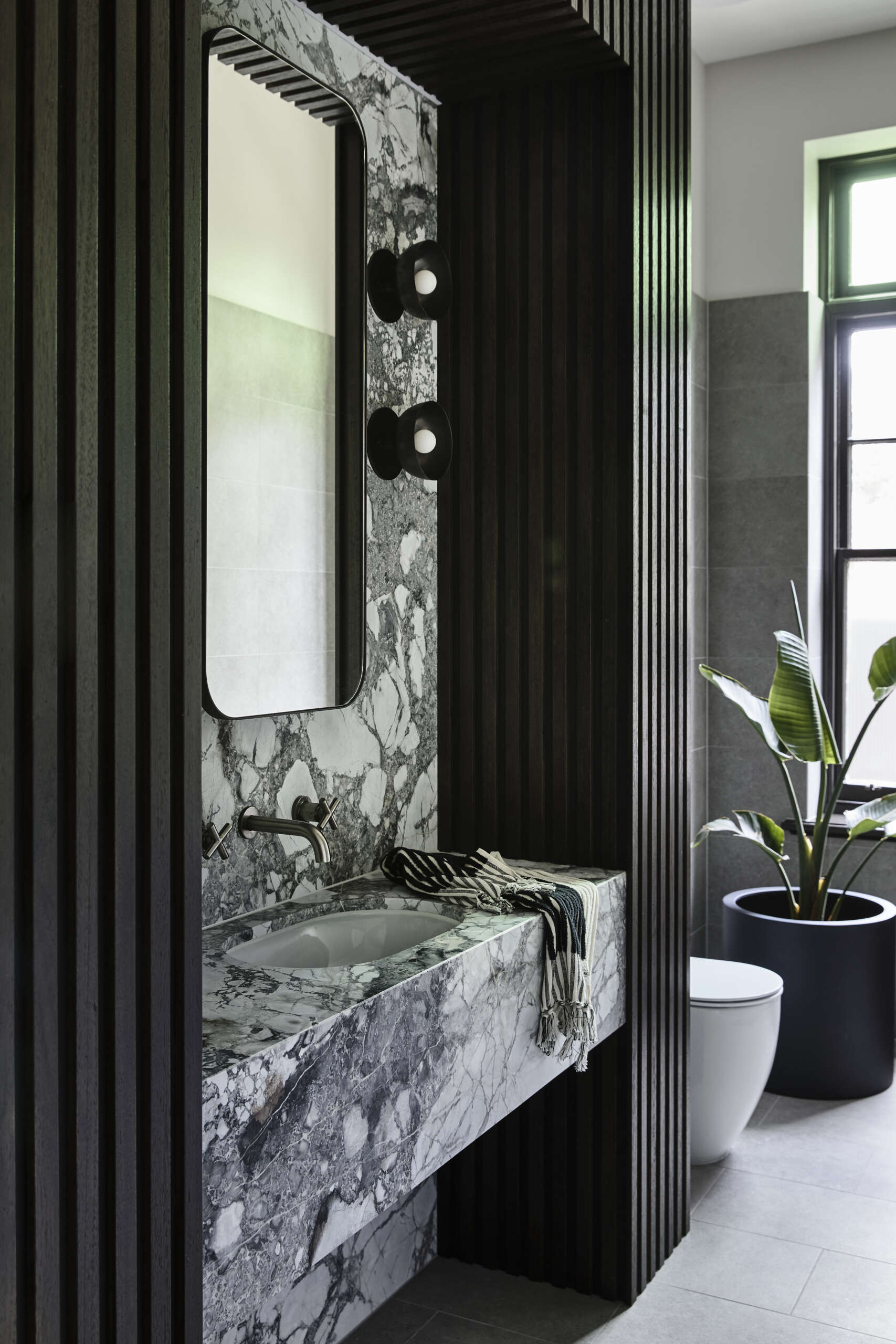 A moody bathroom with a black wood slat wall and sculptural marble vanity. Soft daylight and a leafy plant bring warmth and balance to the darker tones.
