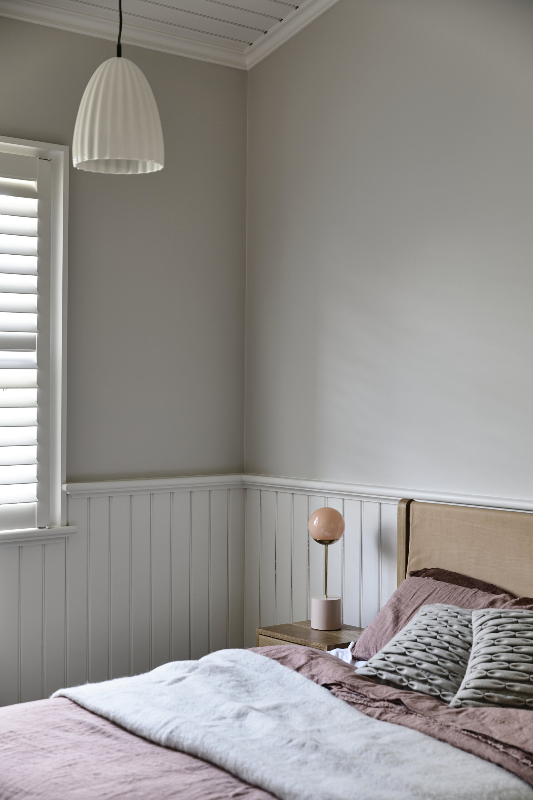 A calming bedroom with soft gray walls, white beadboard, and layered mauve bedding. Natural light, gentle textures, and sculptural lighting keep the space restful and bright.