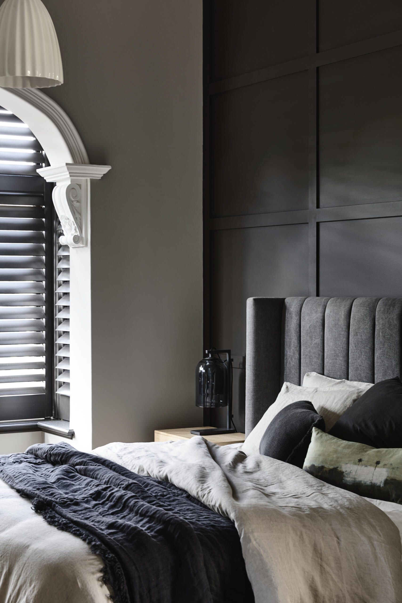 This bedroom embraces a rich, cozy mood with a charcoal paneled wall and plush upholstered headboard. Crisp trim and arched window details balance the darker palette beautifully.