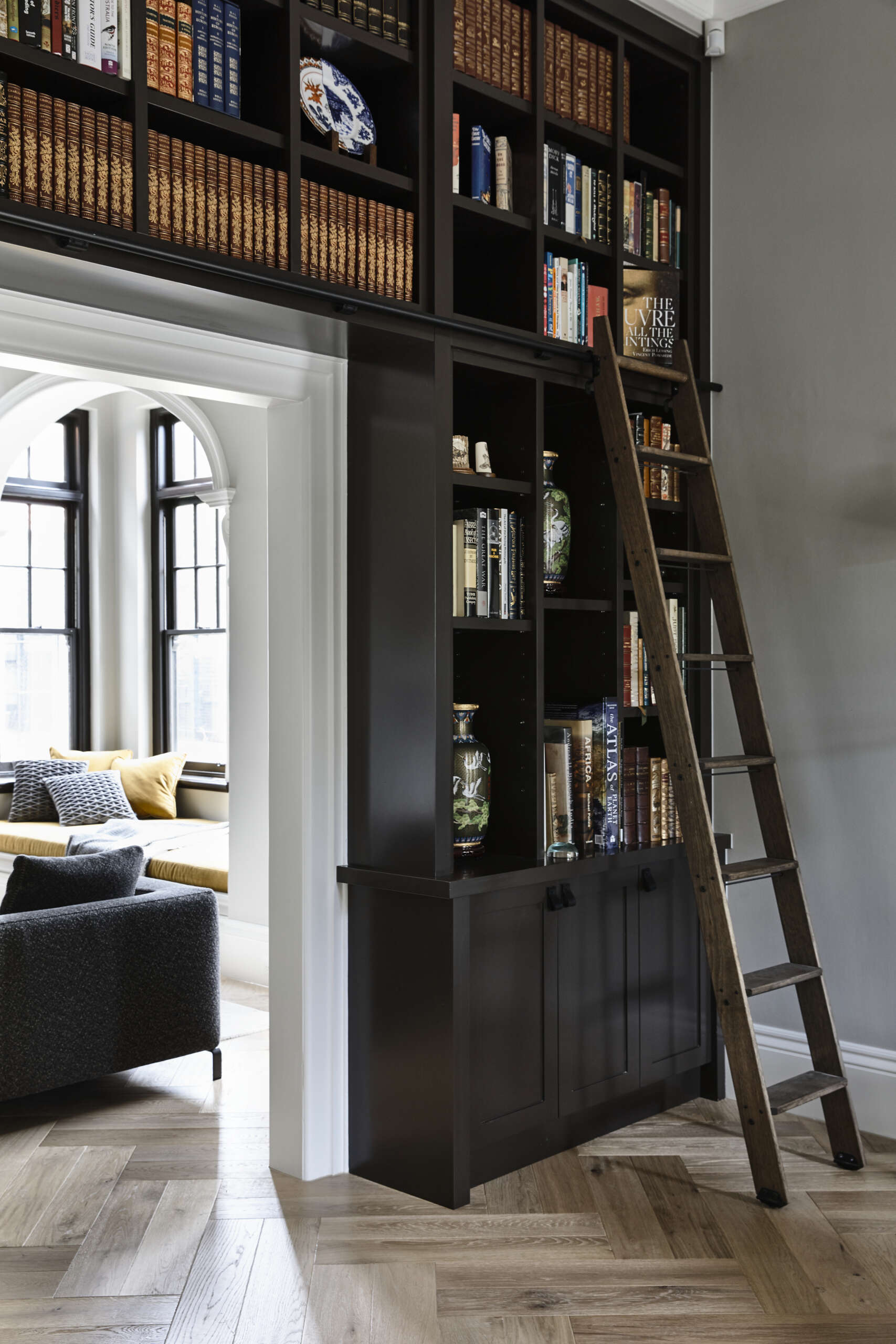 A full-height custom bookshelf with a sliding timber ladder becomes a striking architectural feature. Dark joinery, layered books, and collected objects add depth and character between rooms.