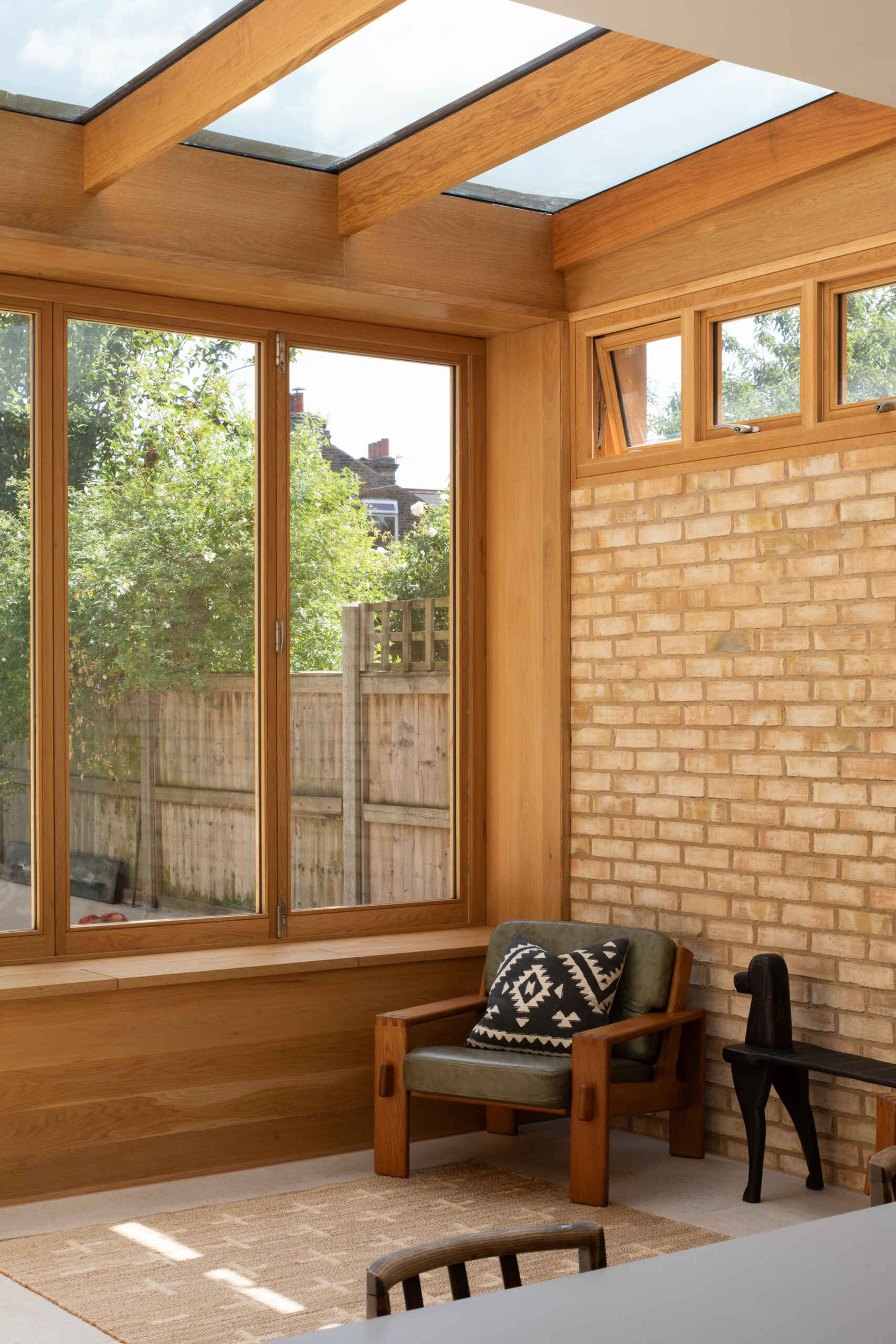 La zona de estar actúa como un suave puente entre la casa y el jardín. Paredes de ladrillo pálido, cálidos marcos de madera y un techo de cristal inundan el espacio de luz durante todo el día. Los suelos de piedra clara mantienen el ambiente luminoso y funcional, mientras que dos cómodas sillas con cojines texturizados invitan a un descanso tranquilo. Una simple consola de madera y flores frescas suavizan la habitación, y una puerta grande permite que la sensación informal del interior se conecte naturalmente con el jardín exterior.