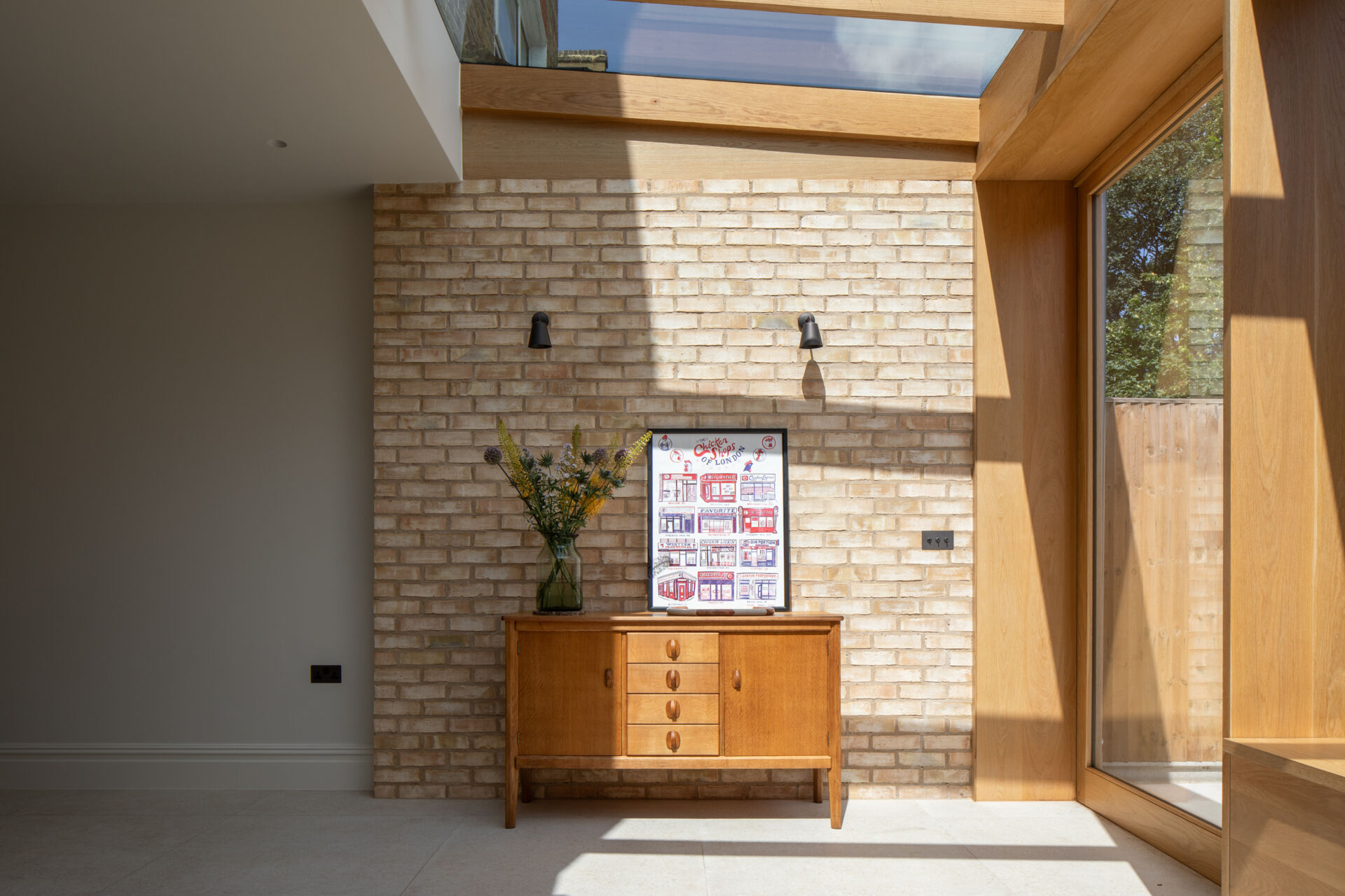 La zona de estar actúa como un suave puente entre la casa y el jardín. Paredes de ladrillo pálido, cálidos marcos de madera y un techo de cristal inundan el espacio de luz durante todo el día. Los suelos de piedra clara mantienen el ambiente luminoso y funcional, mientras que dos cómodas sillas con cojines texturizados invitan a un descanso tranquilo. Una simple consola de madera y flores frescas suavizan la habitación, y una puerta grande permite que la sensación informal del interior se conecte naturalmente con el jardín exterior.