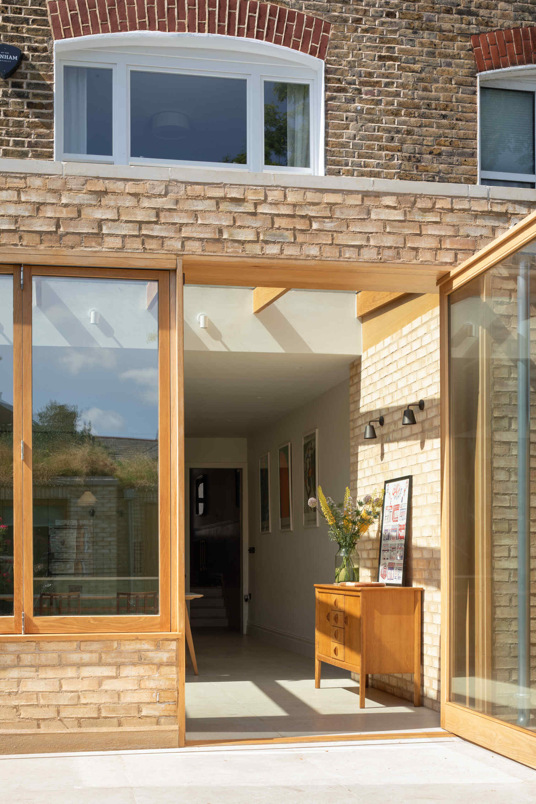 Vista desde el jardín, la ampliación crea un contraste lleno de luz con el clásico ladrillo de la casa. Los cálidos marcos de madera y los amplios paneles de vidrio se abren al patio trasero, permitiendo que la luz del sol fluya libremente a través de los pisos de piedra y hacia la cocina.