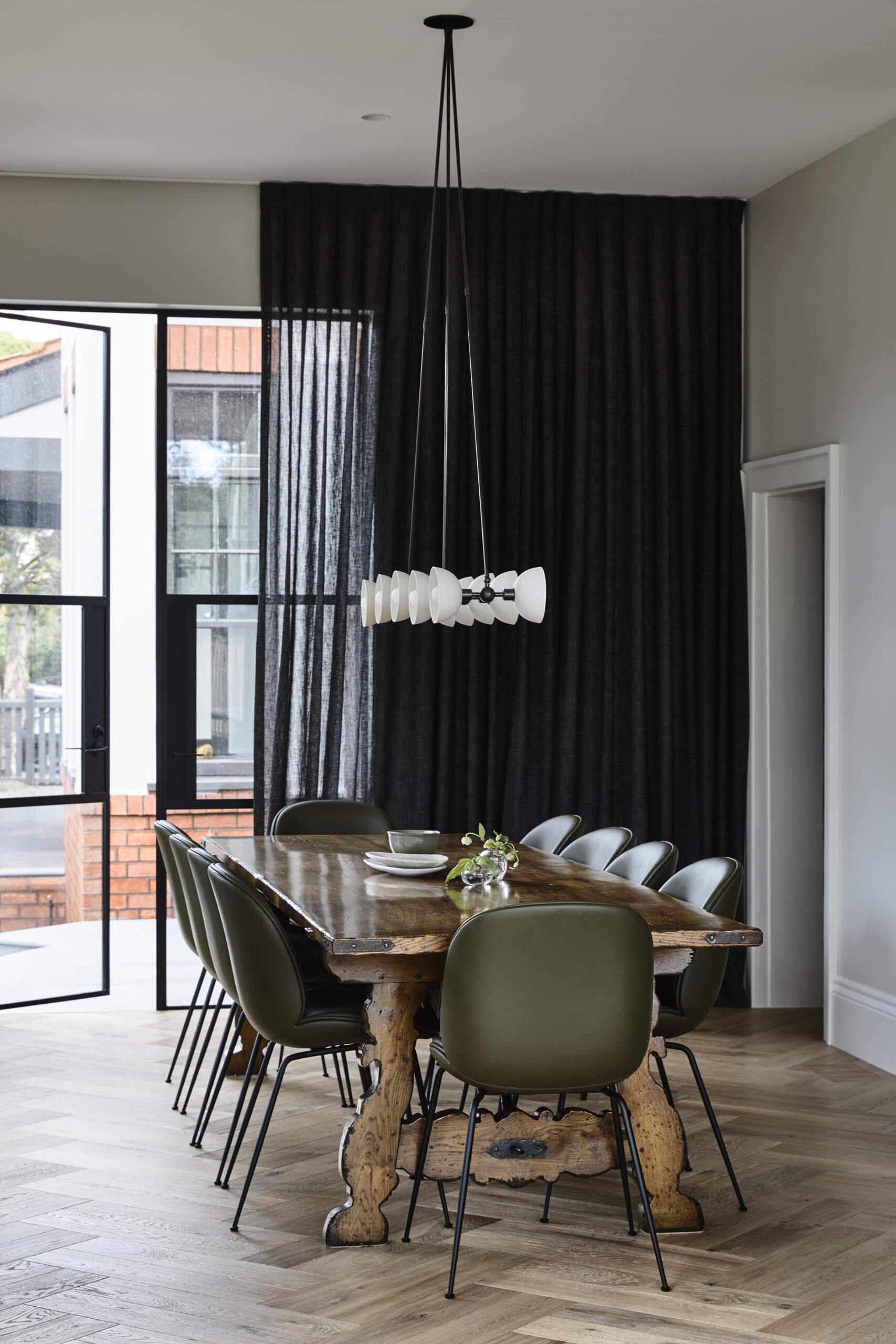This dining room pairs a solid timber table with sculptural lighting and dark curtains for an intimate, modern feel. Steel-framed doors connect the space to the garden and fill it with light.