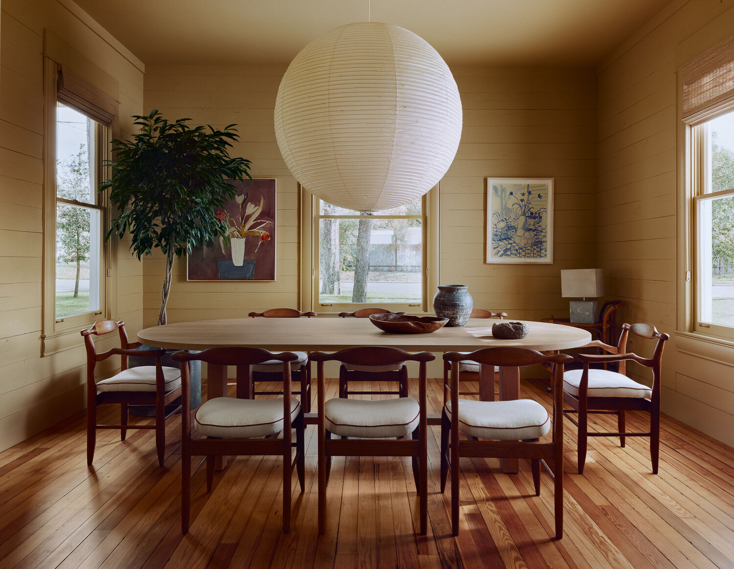 The dining room is described as the heart of the home, with original wood pocket doors creating a striking entry into the room. Original shiplap walls were preserved and painted a soft neutral yellow, while an oval Nickey Kehoe community table anchors the space, designed for large gatherings. Vintage Guillerme et Chambron dining chairs and matching armchairs surround the table, all reupholstered in a cream fabric with rust red trim. An oversized Noguchi pendant casts a warm glow above.
