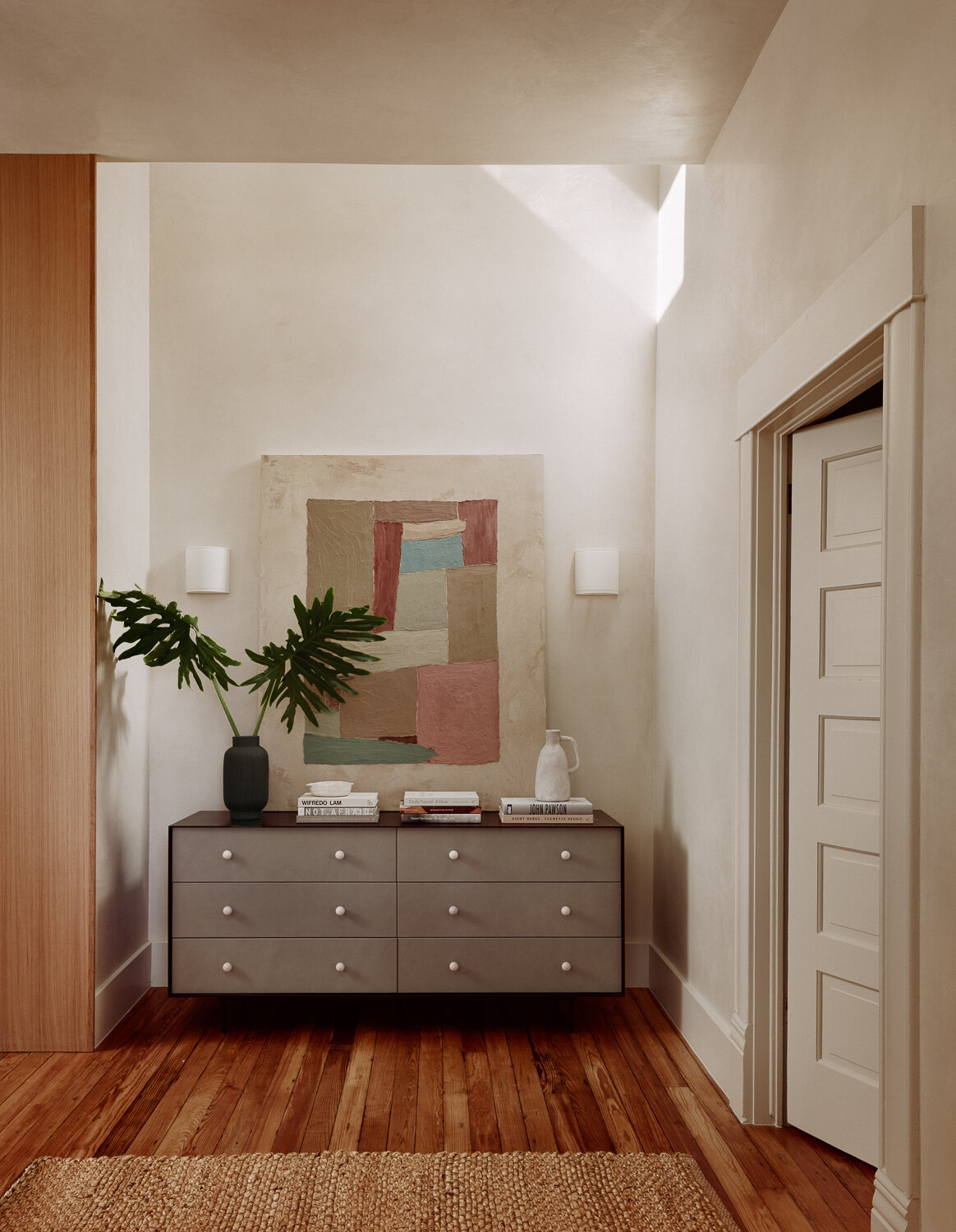 The entryway sets a calm tone from the moment you step inside. Creamy walls and warm wood floors create a light, grounded backdrop, finished with a simple woven rug underfoot. A low dresser keeps storage discreet, while petite white sconces provide gentle, even light. Oversized abstract artwork introduces muted color, balanced by greenery and sculptural accents that make the space feel composed without feeling precious.
