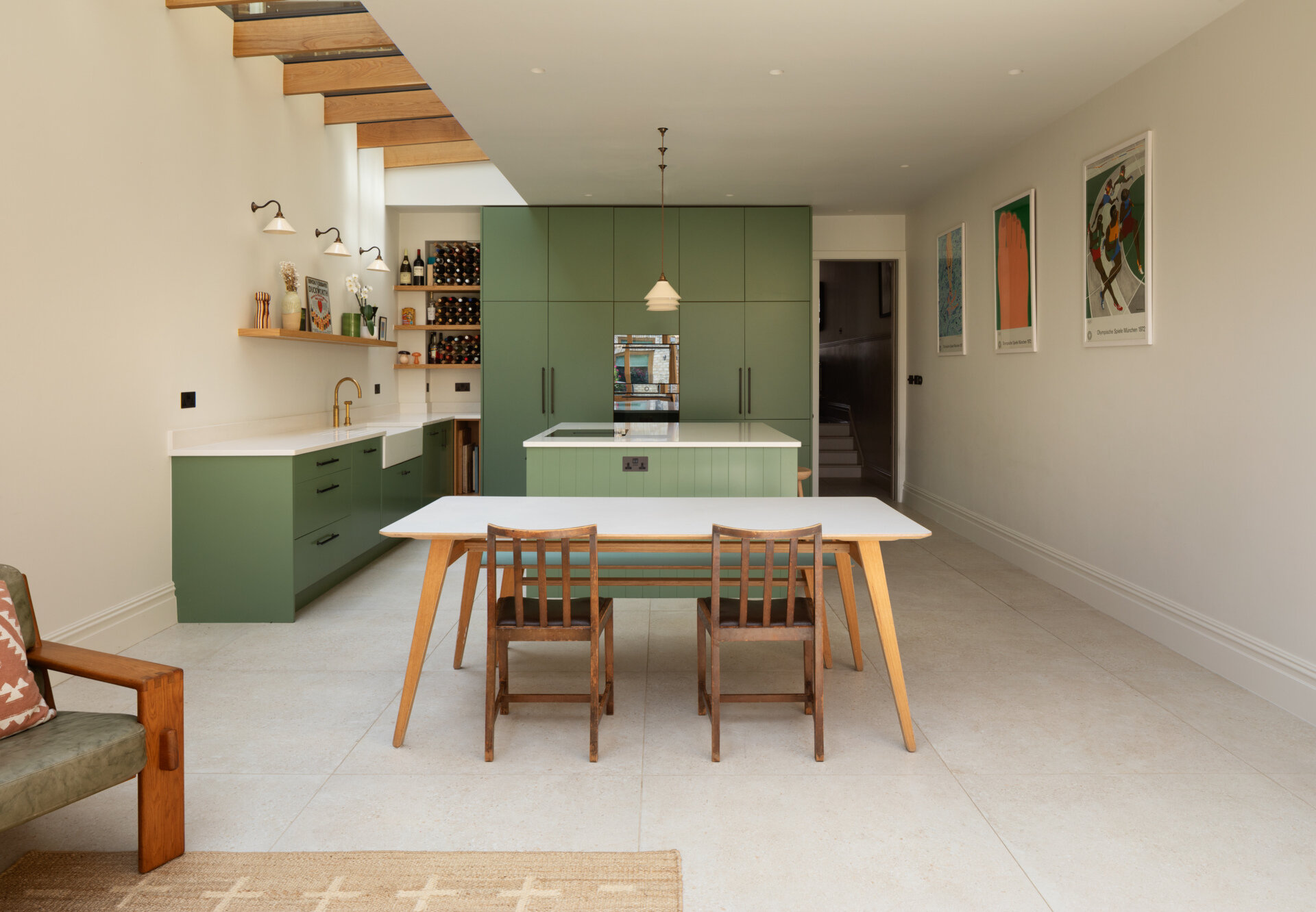 Los gabinetes de color verde salvia suave crean un tono tranquilo y moderno para esta cocina y comedor. Las encimeras de color blanco nítido y los cálidos grifos de latón añaden contraste, mientras que la luz natural desde arriba hace que los pisos de piedra clara se sientan aireados y habitables. Las estanterías de madera abiertas, un rincón para vinos incorporado y una isla estilizada mantienen el espacio organizado y perfecto para cocinar todos los días. Los candelabros y apliques de pared simples, junto con el divertido arte mural, hacen una declaración sin agregar desorden.