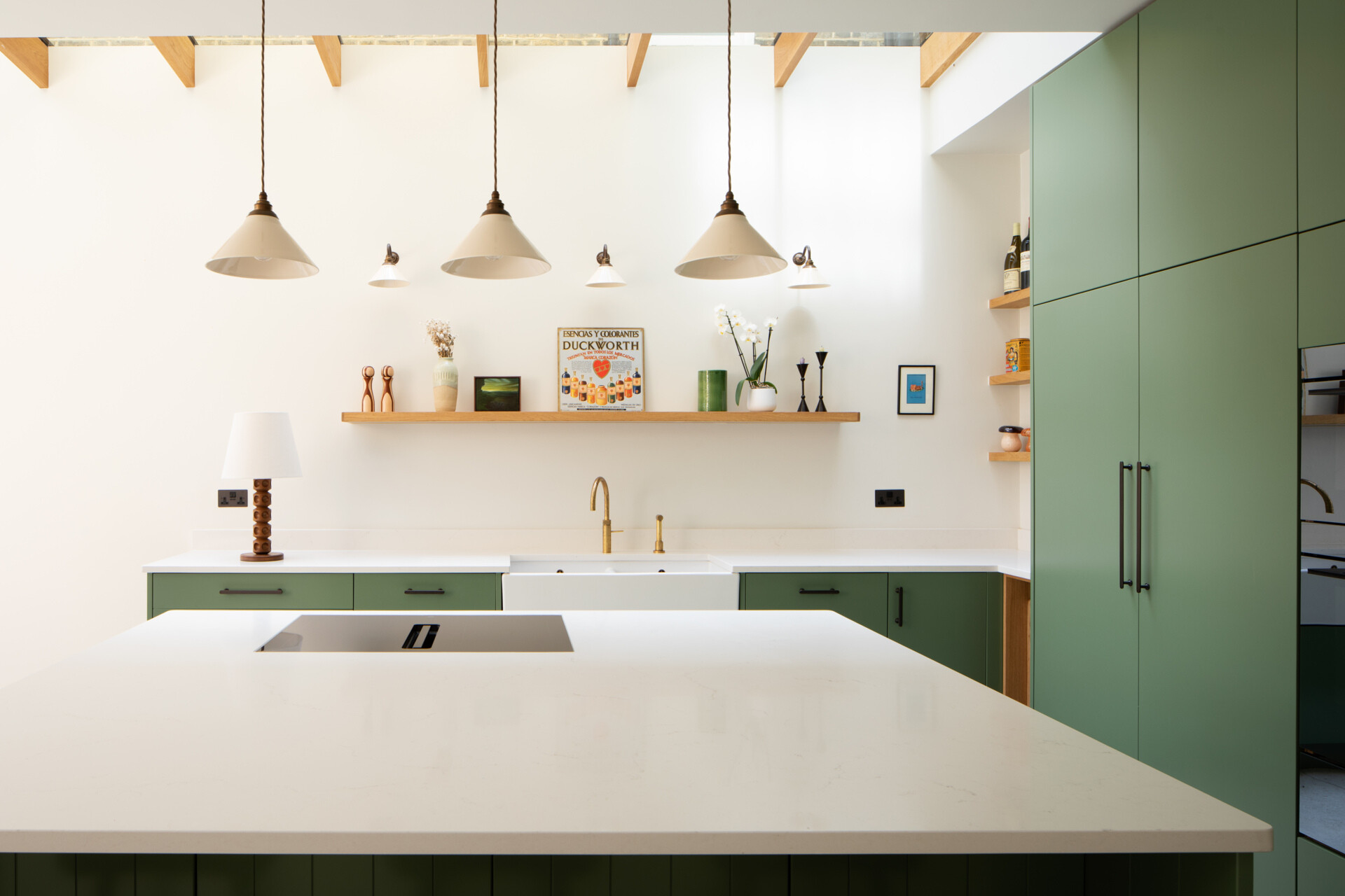 Los gabinetes de color verde salvia suave crean un tono tranquilo y moderno para esta cocina y comedor. Las encimeras de color blanco nítido y los cálidos grifos de latón añaden contraste, mientras que la luz natural desde arriba hace que los pisos de piedra clara se sientan aireados y habitables. Las estanterías de madera abiertas, un rincón para vinos incorporado y una isla estilizada mantienen el espacio organizado y perfecto para cocinar todos los días. Los candelabros y apliques de pared simples, junto con el divertido arte mural, hacen una declaración sin agregar desorden.