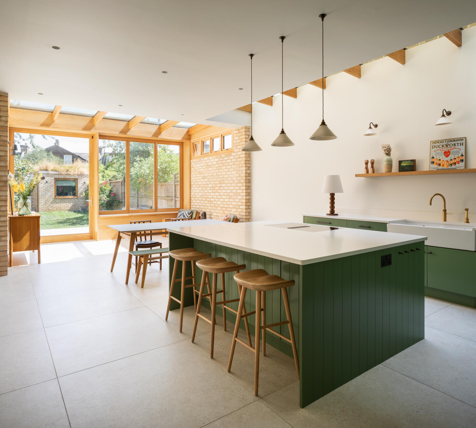 Los gabinetes de color verde salvia suave crean un tono tranquilo y moderno para esta cocina y comedor. Las encimeras de color blanco nítido y los cálidos grifos de latón añaden contraste, mientras que la luz natural desde arriba hace que los pisos de piedra clara se sientan aireados y habitables. Las estanterías de madera abiertas, un rincón para vinos incorporado y una isla estilizada mantienen el espacio organizado y perfecto para cocinar todos los días. Los candelabros y apliques de pared simples, junto con el divertido arte mural, hacen una declaración sin agregar desorden.